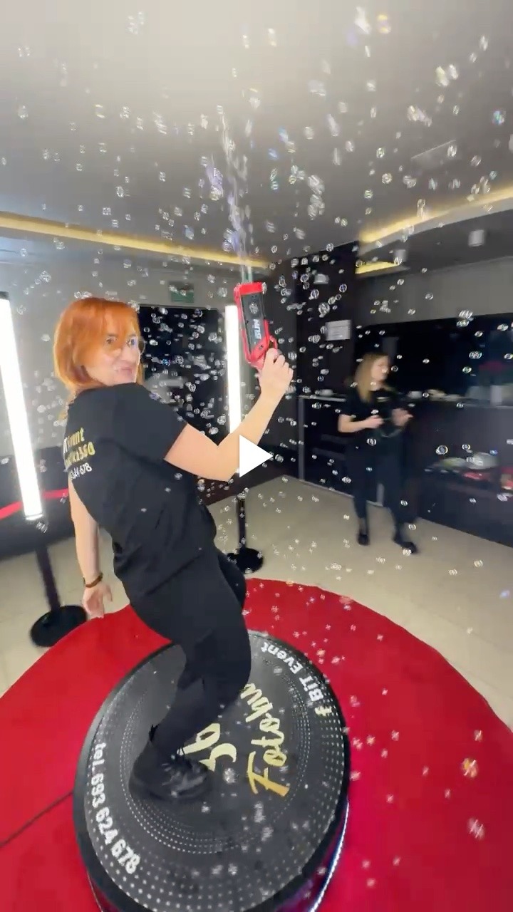 Kobieta z czerwonymi włosami i czarnym T-shirtem z logo firmy stoi na obrotowej platformie z czerwonym dywanem, trzymając w ręku pistolet do robienia baniek mydlanych, w tle widać drugą osobę...