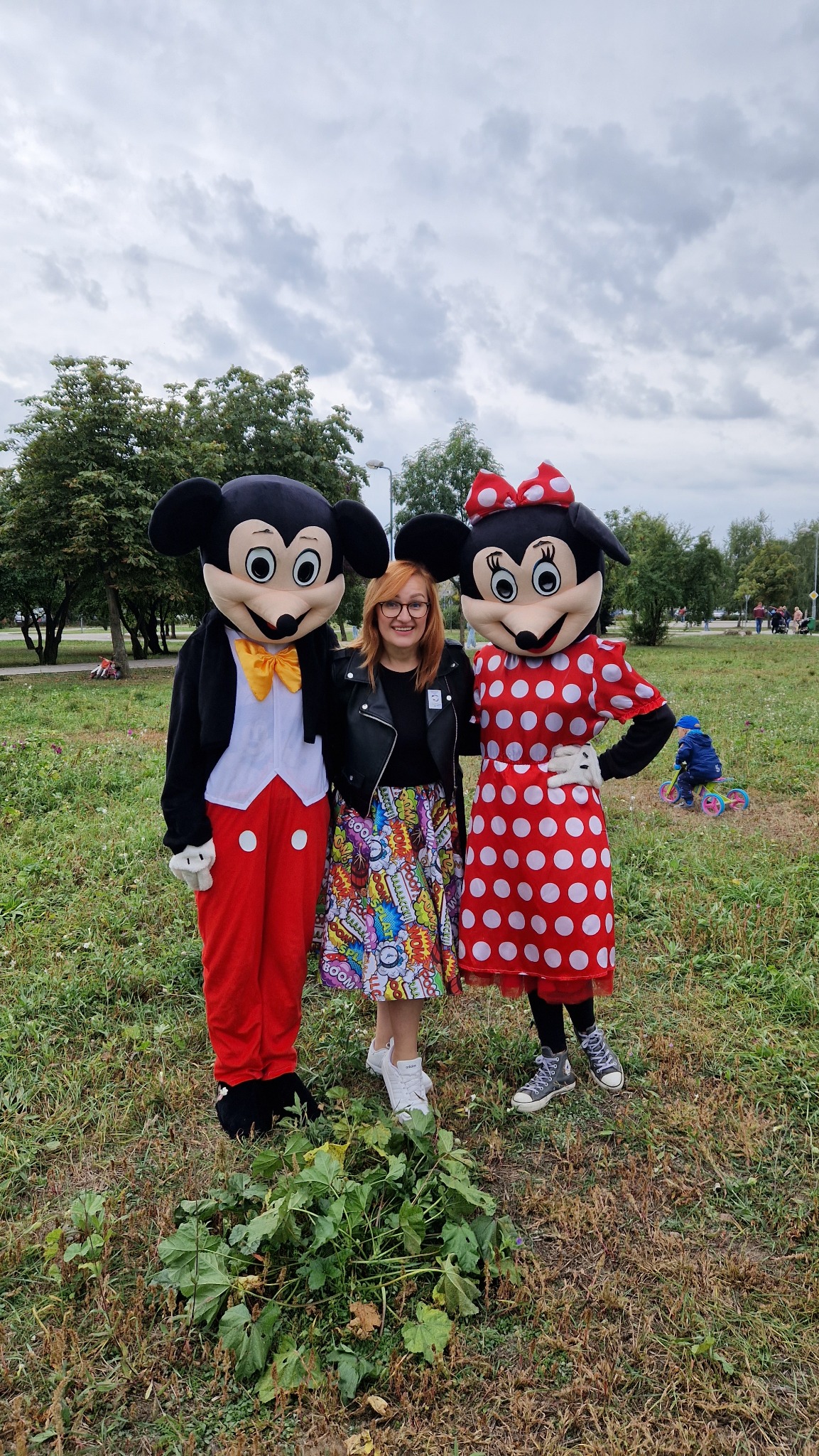 Kobieta w okularach i czarnej kurtce pozuje z postaciami w przebraniach Myszki Miki i Minnie na trawniku, w tle widoczny park i bawiące się dzieci.
