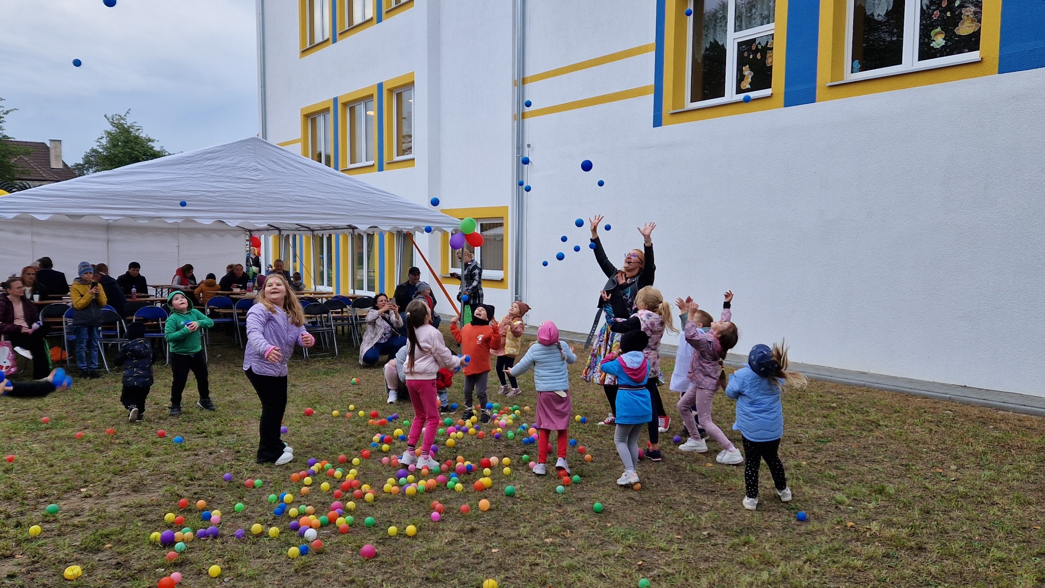 Dzieci bawią się kolorowymi piłeczkami na trawie, animator w okularach rzuca niebieskie piłki do góry, w tle biały budynek z żółto-niebieskimi obramowaniami okien i biały namiot z siedzącymi osobami.