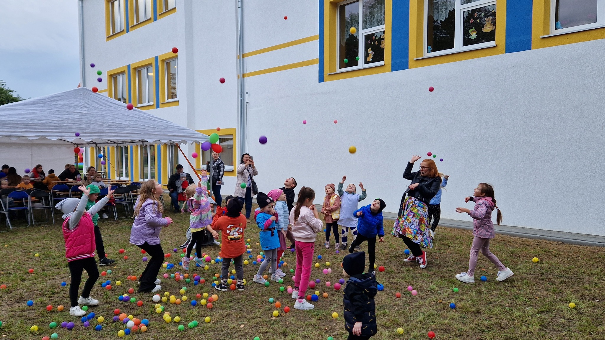 Grupa dzieci bawi się kolorowymi piłkami na trawie przed budynkiem, kobieta w czarnej kurtce i wzorzystej spódnicy rzuca piłki do góry, w tle biały namiot i osoby siedzące przy stołach.