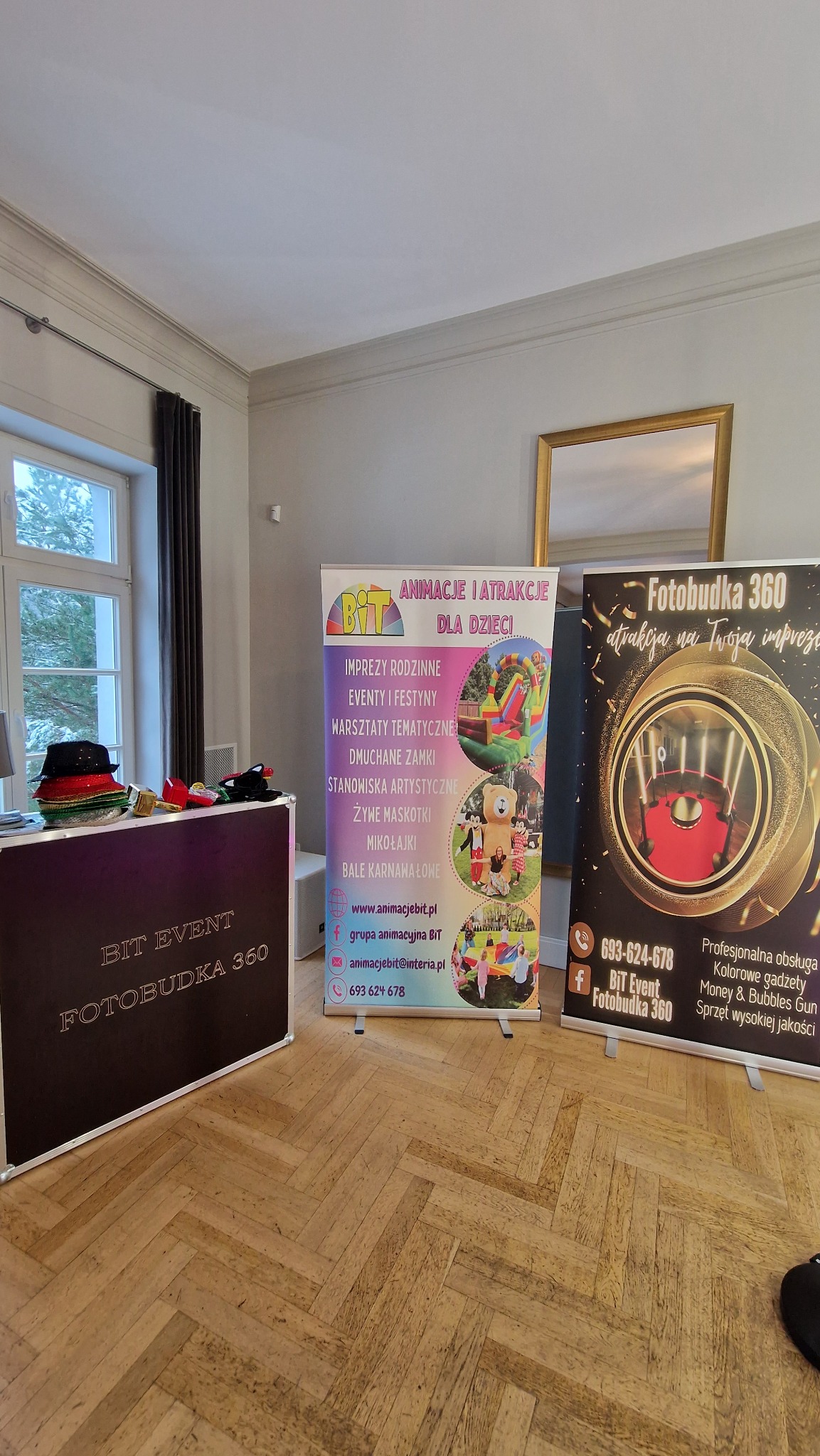 Wnętrze pomieszczenia z parkietem, gdzie ustawiono stoisko z napisem 'Bit Event Fotobudka 360' oraz dwa roll-upy reklamujące animacje dla dzieci i fotobudkę na imprezy.