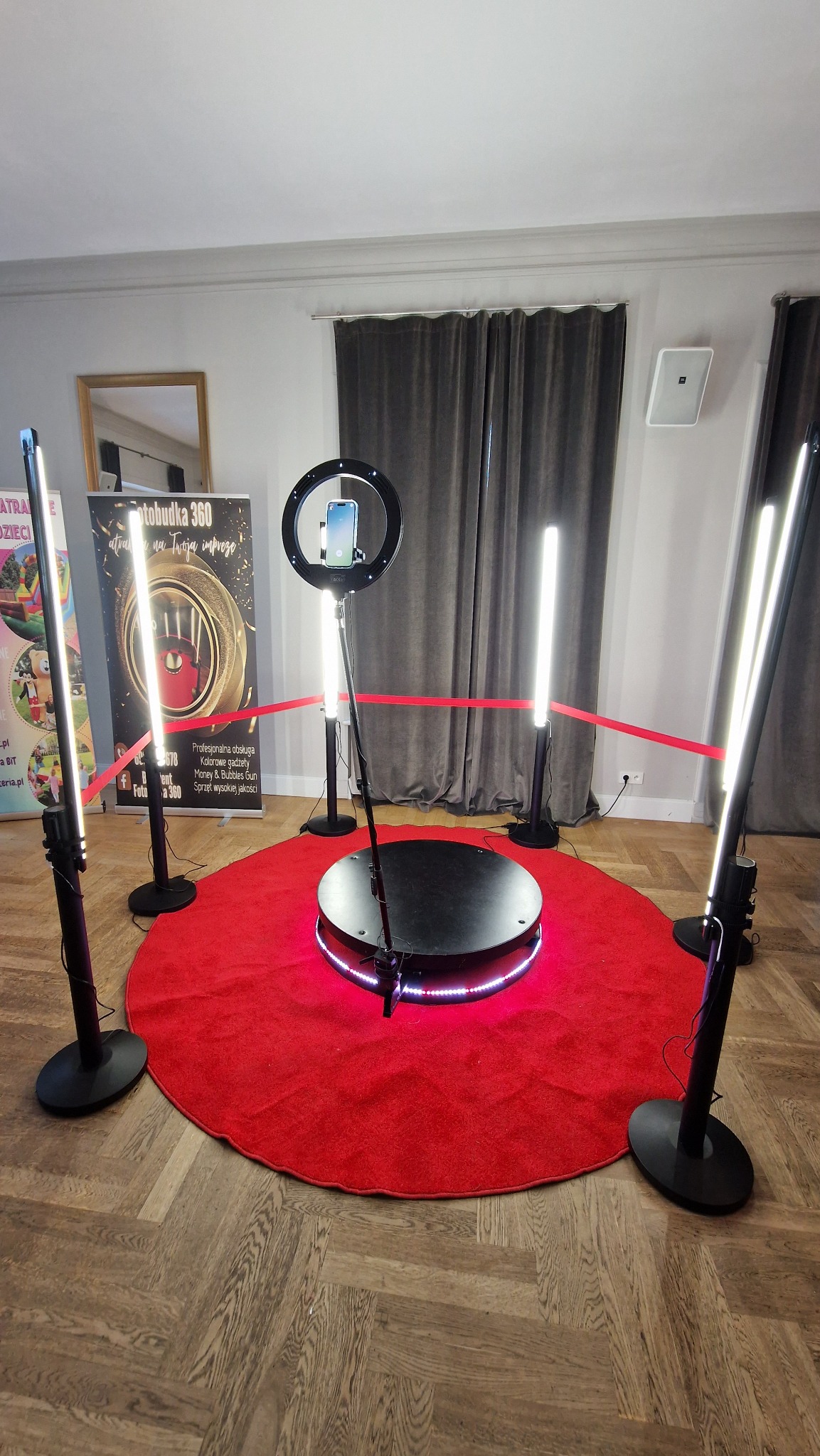 Oświetlona platforma obrotowa 360 stopni z telefonem w uchwycie, otoczona czerwonym dywanem i lampami LED, zabezpieczona czerwoną taśmą.