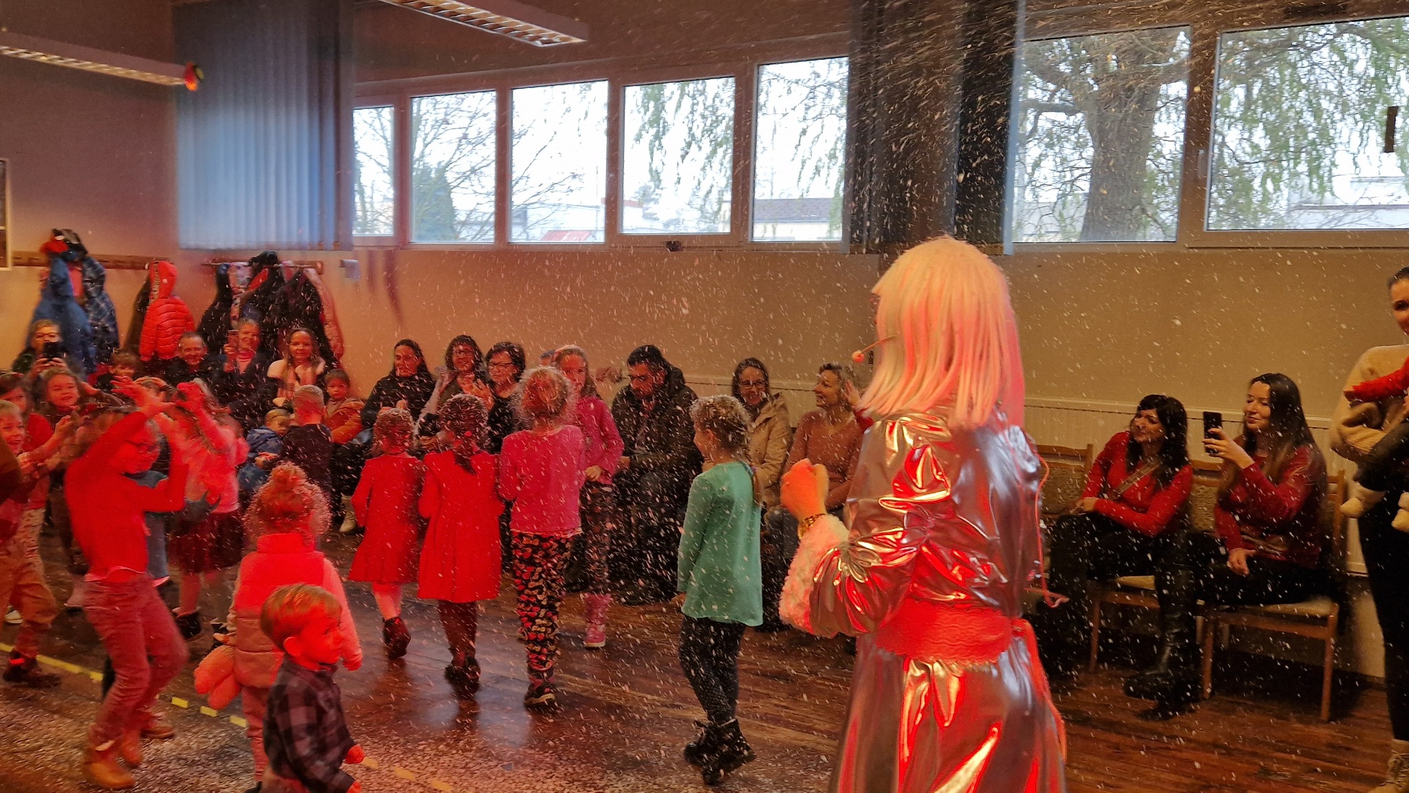 Animator w srebrnym kostiumie i białej peruce prowadzi zabawę z dziećmi, sztuczny śnieg w powietrzu, rodzice obserwują z tyłu, widok zza animatora.