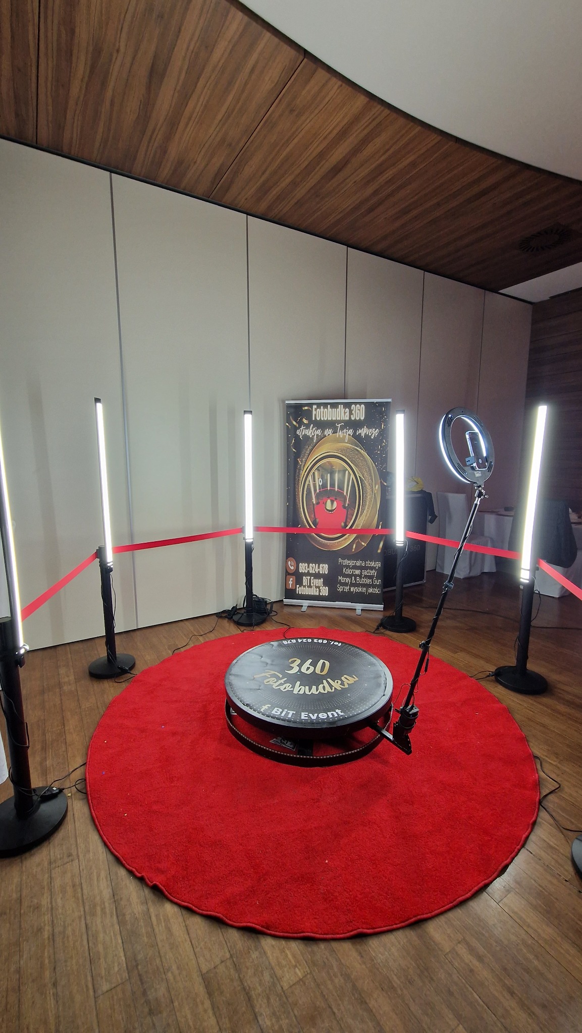 Okrągła platforma 360 z napisem 'Fotobudka' na czerwonym dywanie, otoczona lampami LED i taśmą, z banerem reklamowym w tle