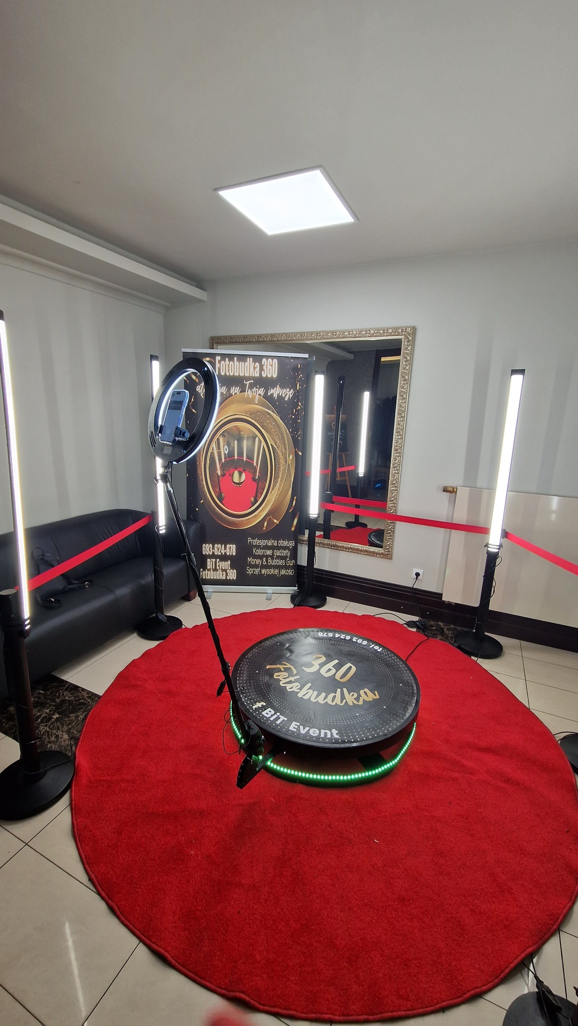 Gotowa do użycia fotobudka 360 na czerwonym dywanie, z oświetleniem LED, telefonem zamocowanym na statywie i banerem reklamowym w tle.