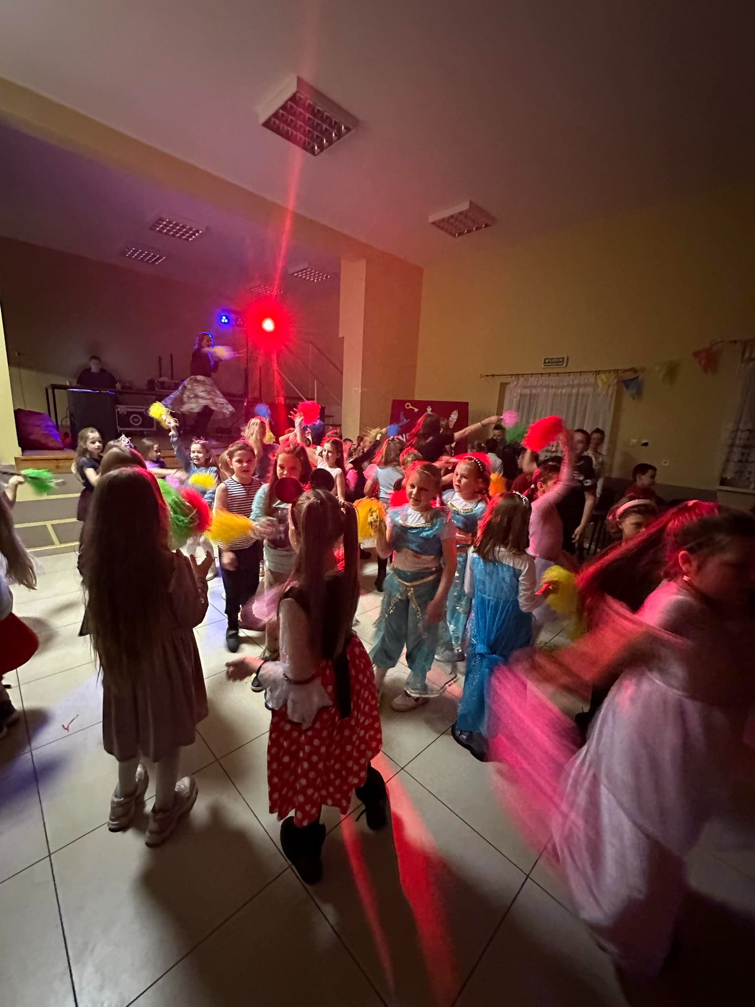 Grupa dzieci w kostiumach karnawałowych tańczy w sali z czerwonym oświetleniem, widoczny DJ na podwyższeniu.
