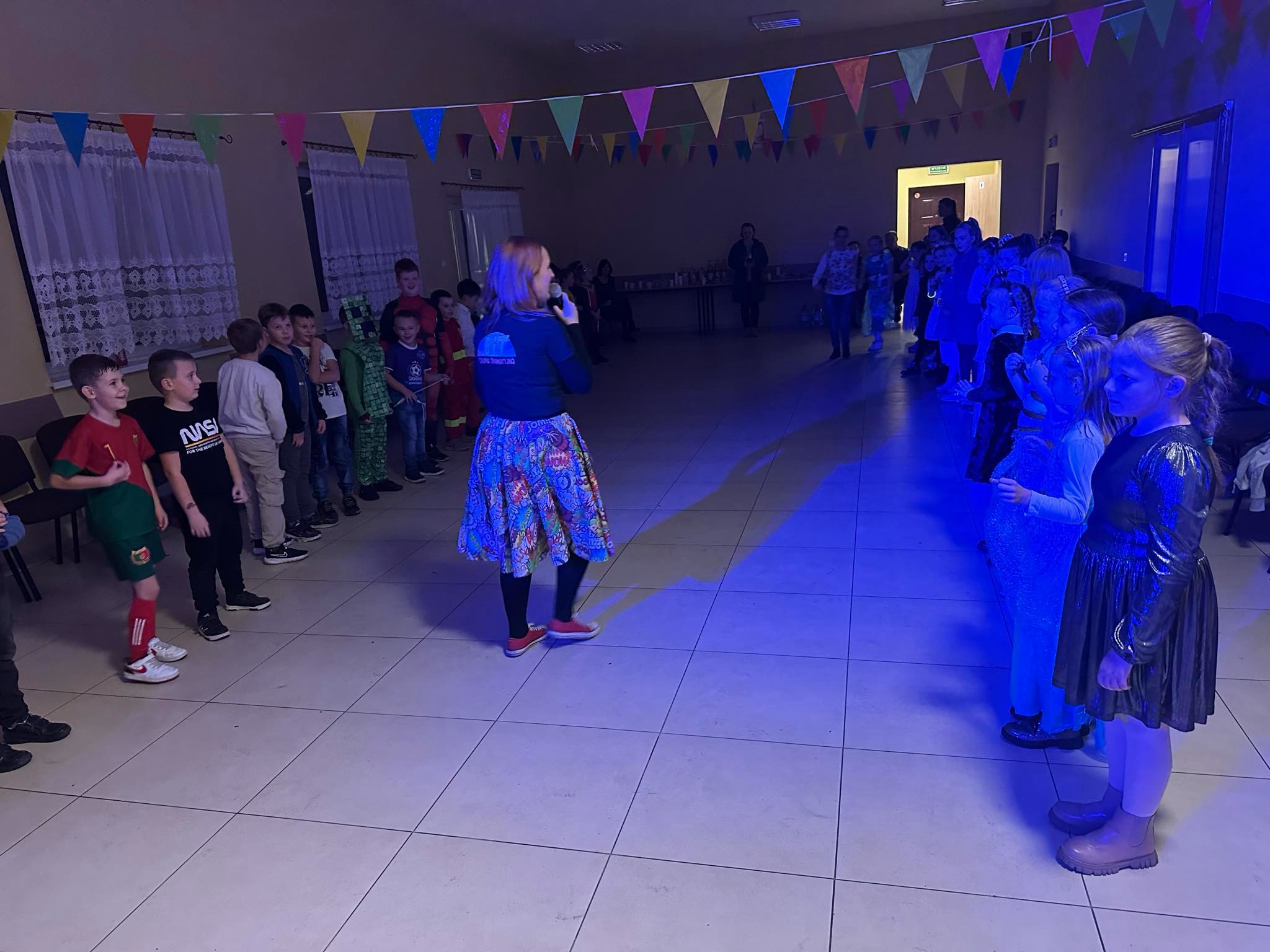 Kobieta z mikrofonem w kolorowej spódnicy prowadzi zabawę dla grupy dzieci w sali udekorowanej kolorowymi chorągiewkami.