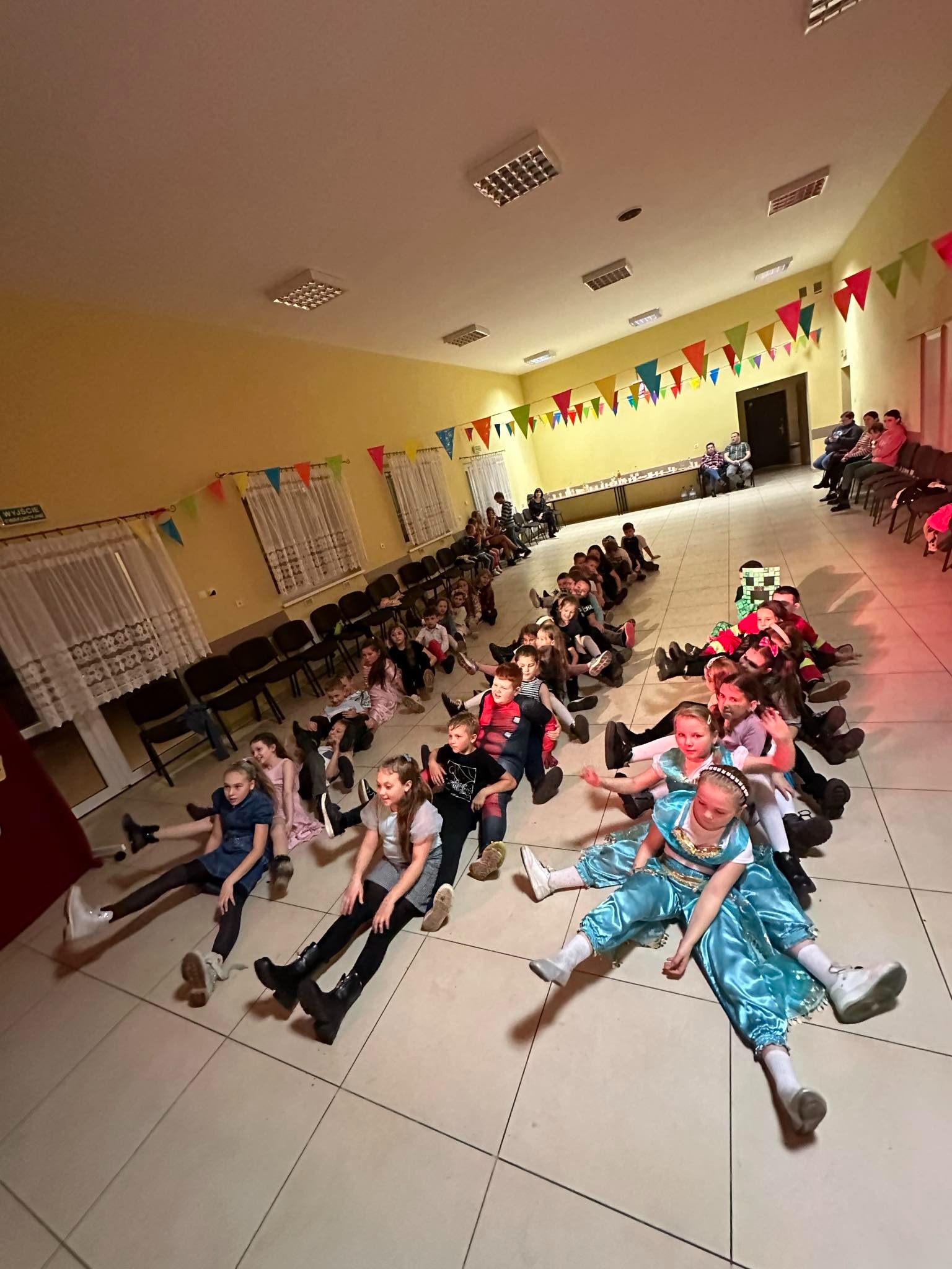 Grupa dzieci w kostiumach siedzi na podłodze w rzędzie podczas animacji w Gołańczy, w tle widoczni rodzice i kolorowe girlandy.