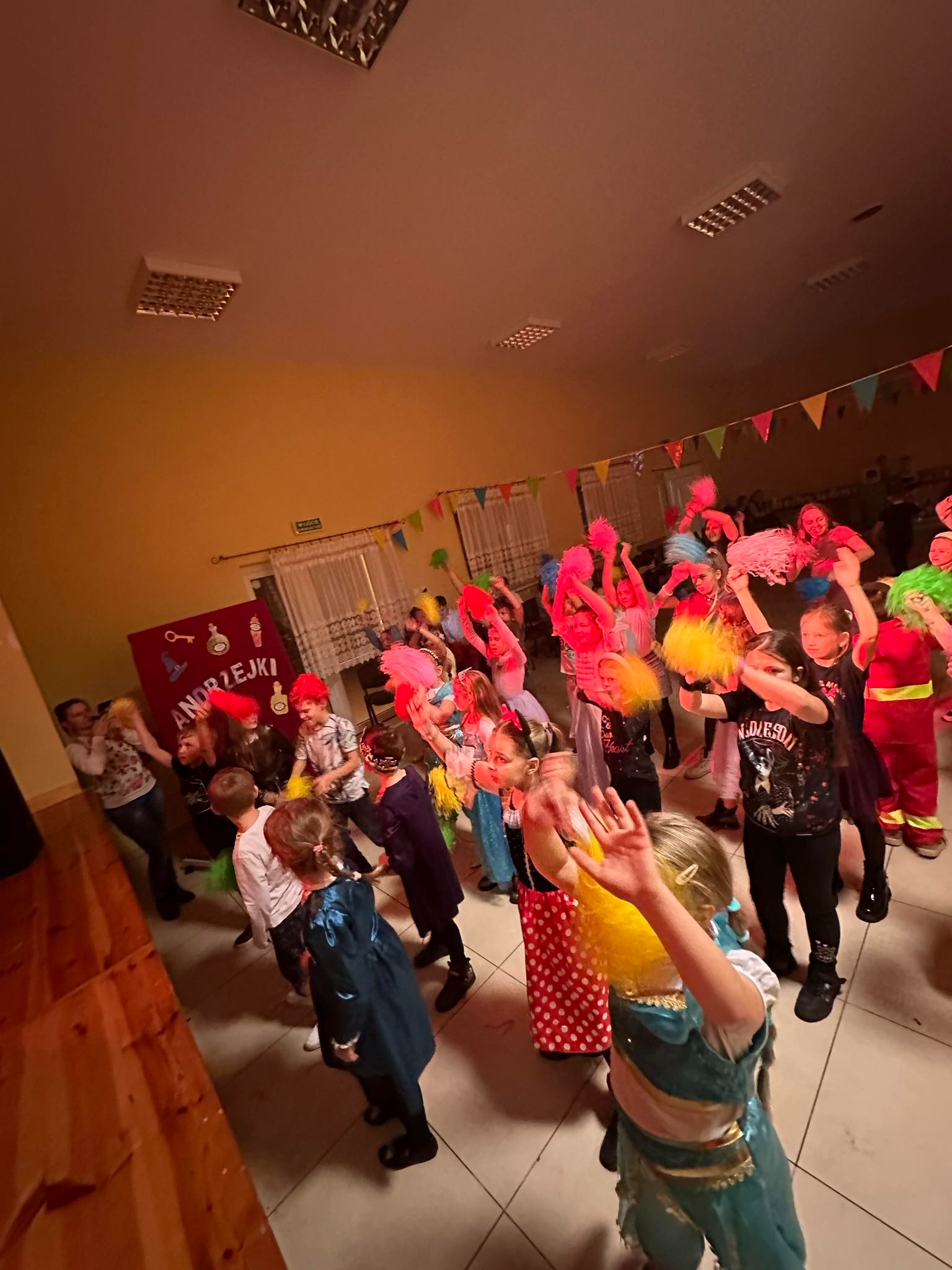 Grupa dzieci w kolorowych przebraniach, z pomponami w rękach, tańczy na tle dekoracji Andrzejkowych w sali z girlandami.