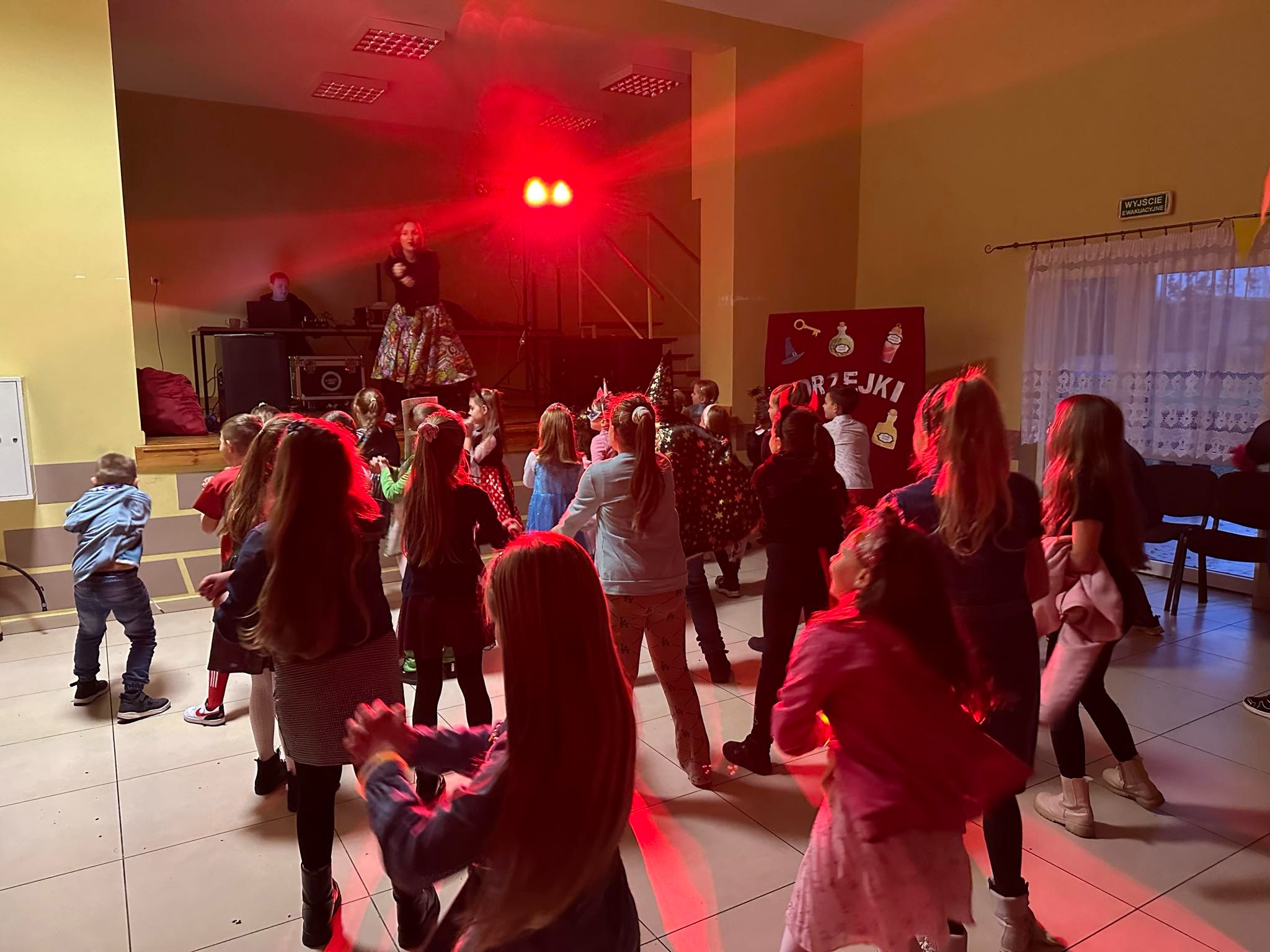 Dzieci tańczą na sali podczas imprezy, oświetlenie czerwone, na scenie kobieta w kwiecistej spódnicy i mężczyzna za konsoletą.