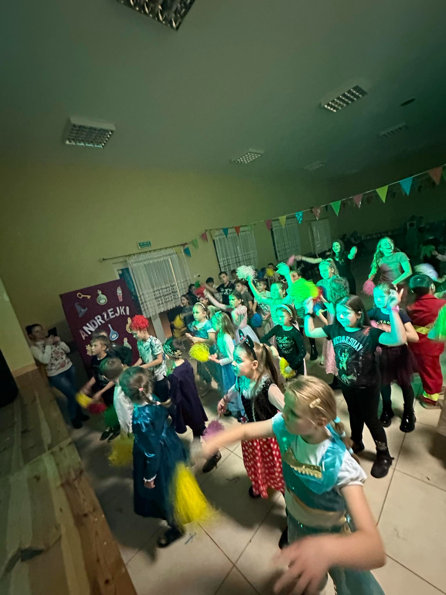 Sala z bawiącymi się dziećmi podczas imprezy Andrzejkowej, dekoracje z flagietek, zielone światło, dzieci w przebraniach i z pomponami w dłoniach, tablica z napisem 'Andrzejki'.