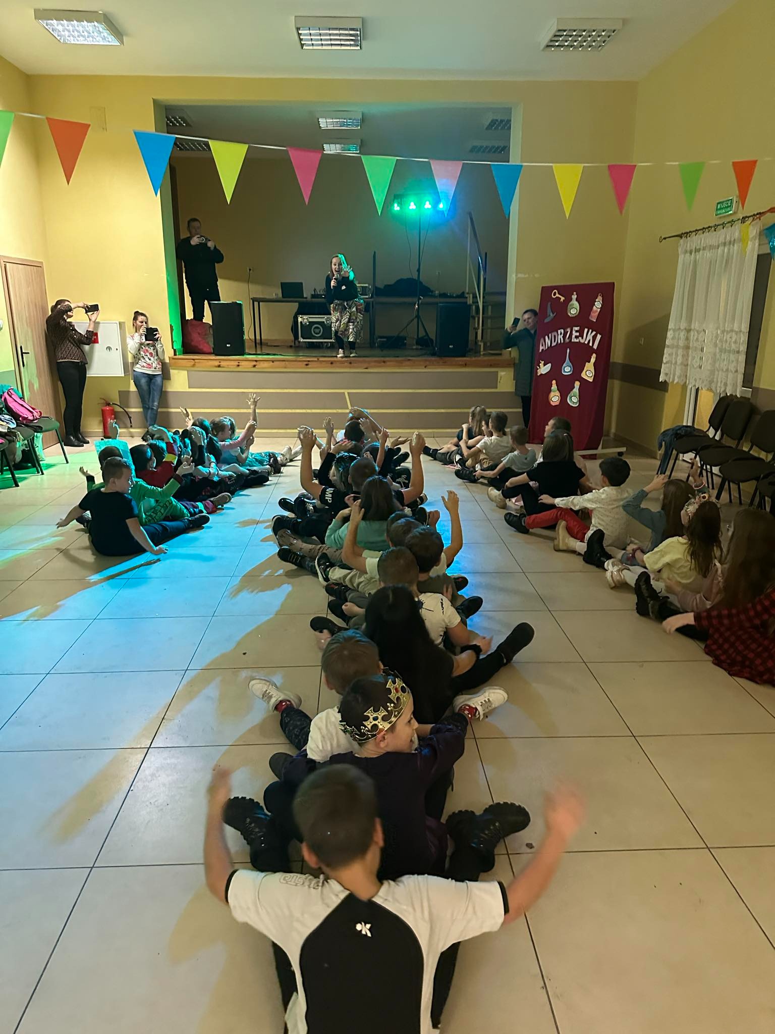 Dzieci siedzące rzędami na podłodze sali, z podniesionymi rękami, obserwują scenę z wokalistką i muzykami podczas imprezy Andrzejkowej, dekoracje z kolorowych trójkątnych flag wiszą pod sufitem.