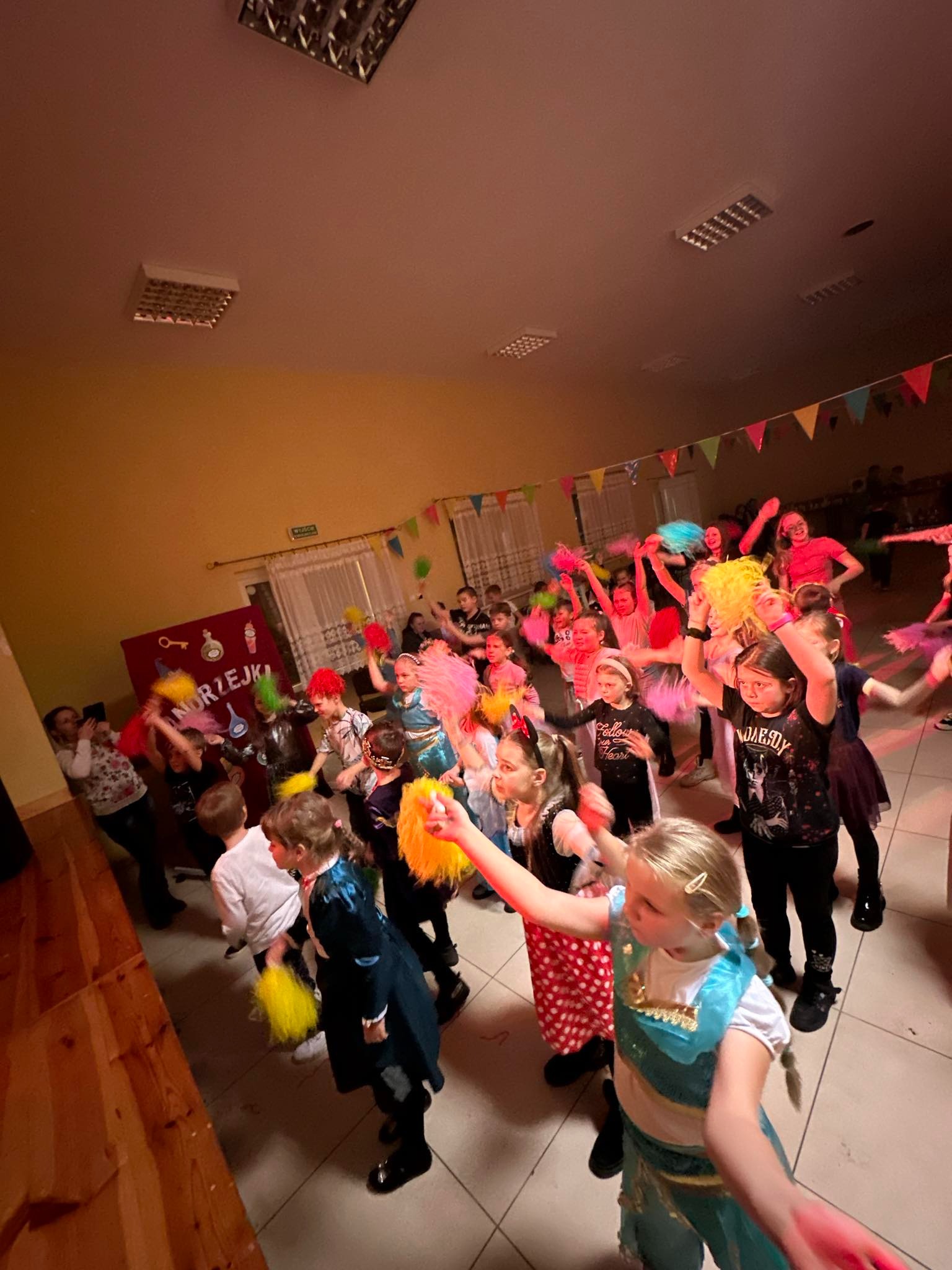 Grupa dzieci w kostiumach i perukach, trzymających kolorowe pompony, podczas występu na tle dekoracji z flagietkami i napisem na czerwonym tle.