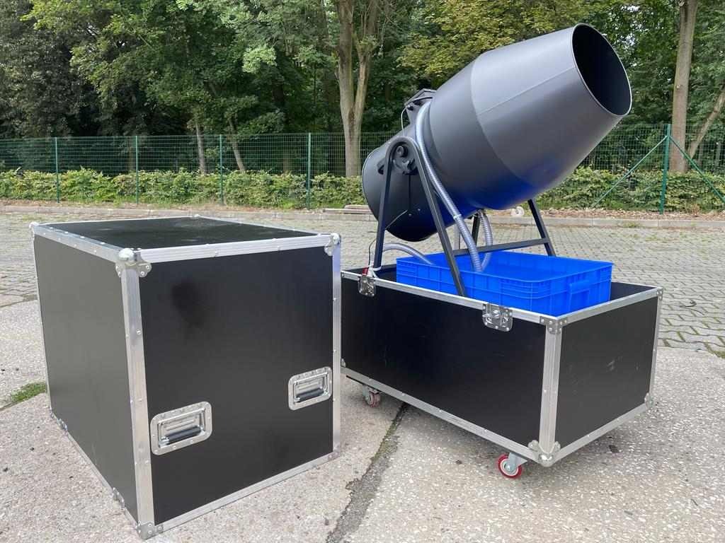 Profesjonalna wytwornica baniek mydlanych w czarnej obudowie transportowej z niebieskim pojemnikiem na płyn, ustawiona na zewnątrz na kostce brukowej, obok pusta skrzynia transportowa.