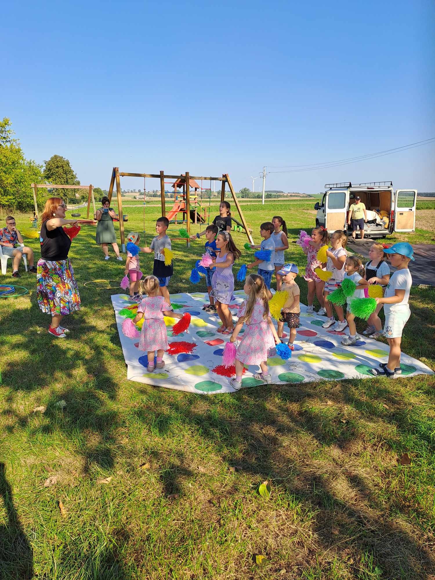Animator prowadzi grupę dzieci w grze na kolorowej macie na trawie; w tle huśtawka, bus i pole.