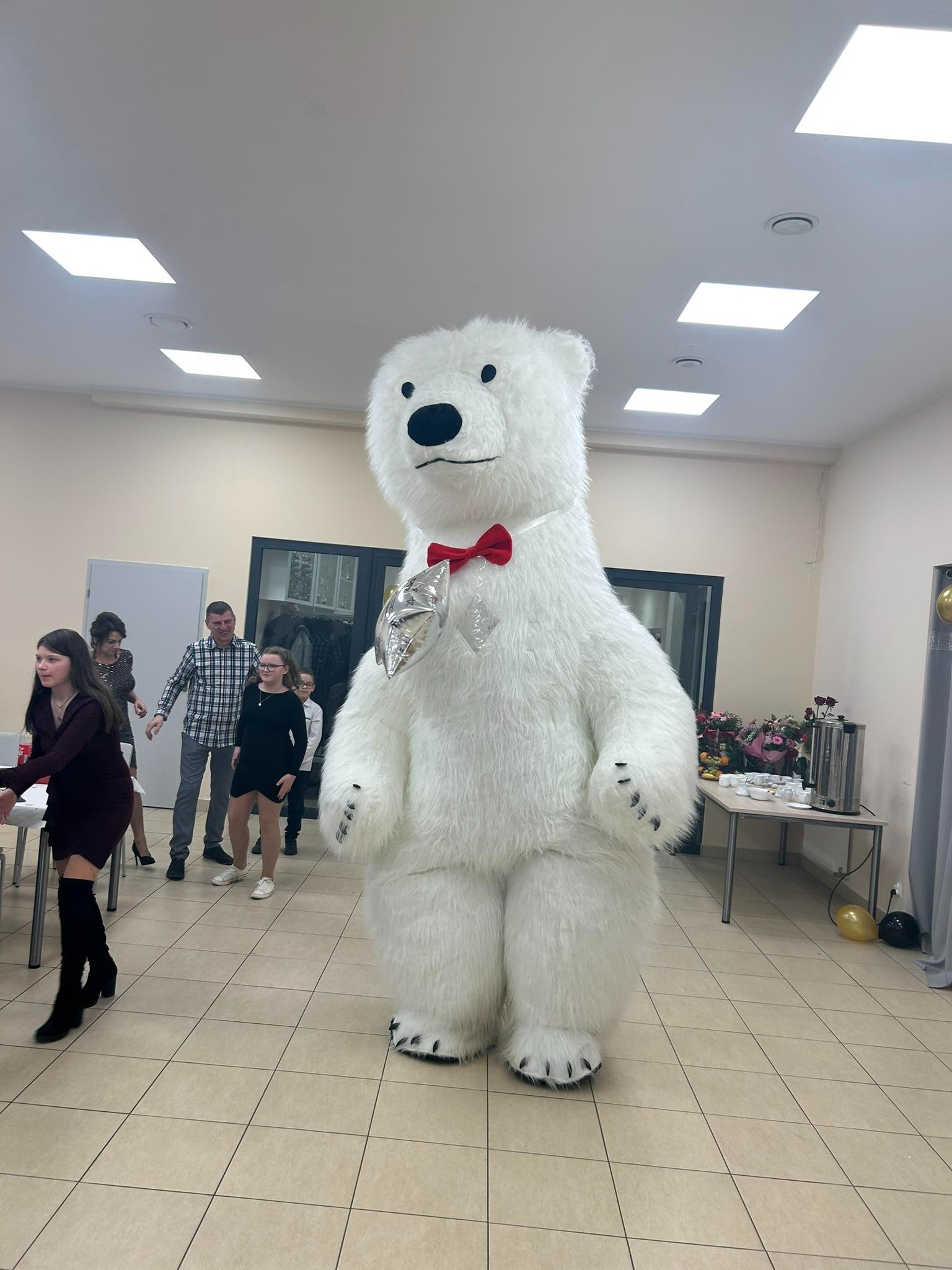 Duży, biały miś w kostiumie z czerwoną muszką i srebrnym balonem w kształcie serca stoi na kafelkach, w tle grupa ludzi i stół z kwiatami.