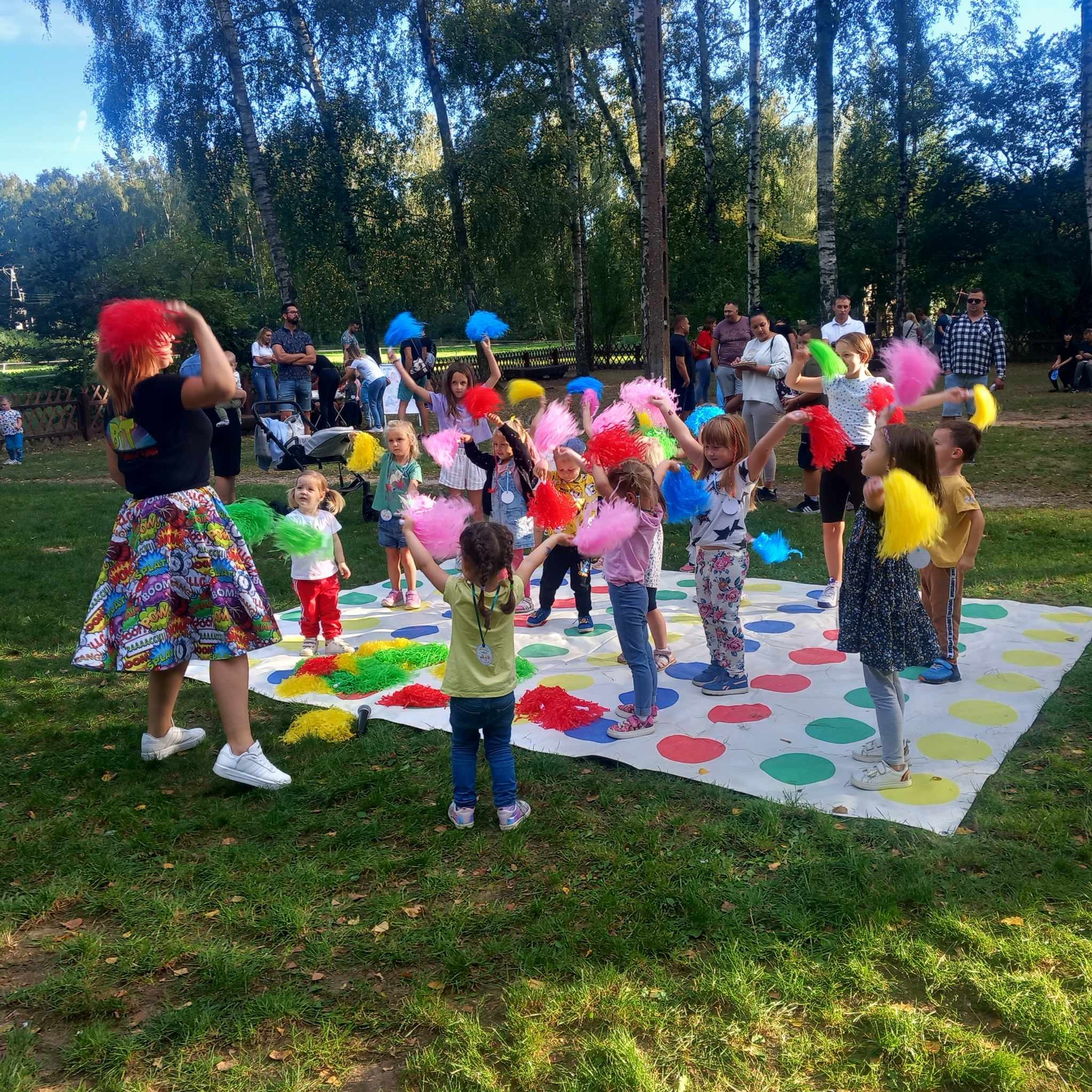 Grupa dzieci z kolorowymi pomponami na trawie, grających w Twistera pod okiem animatorki w spódnicy w komiksowe wzory, w tle park i obserwujący rodzice.