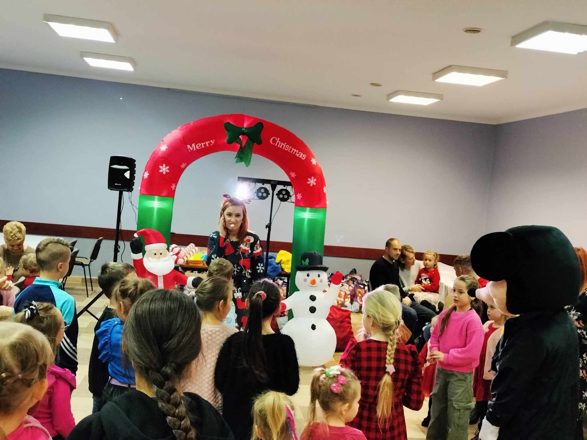 Kobieta z pomalowaną twarzą w świątecznym swetrze prowadzi animację dla grupy dzieci, w tle dmuchany łuk z napisem Merry Christmas, dmuchany bałwan i Święty Mikołaj, oraz osoba w stroju Myszki Miki.