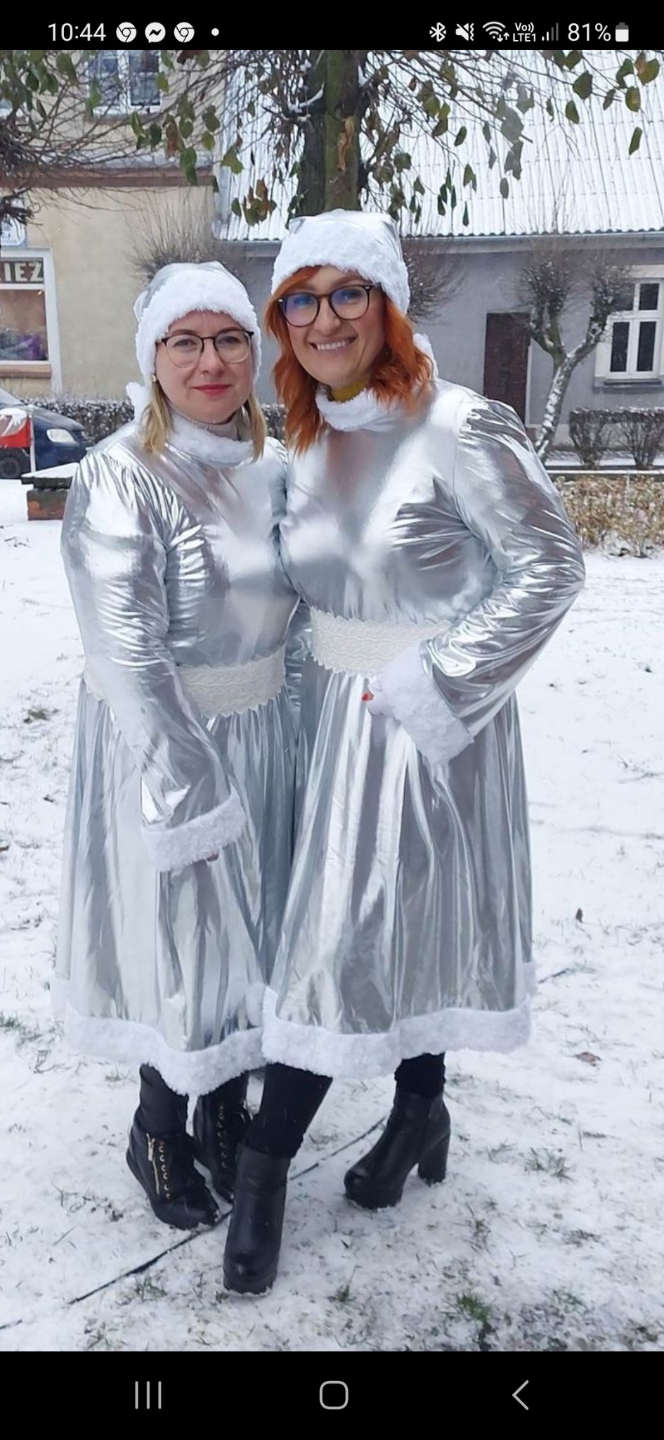 Dwie kobiety w srebrnych kostiumach z białym futerkiem i czapkach Mikołaja stoją na śniegu przed budynkiem.