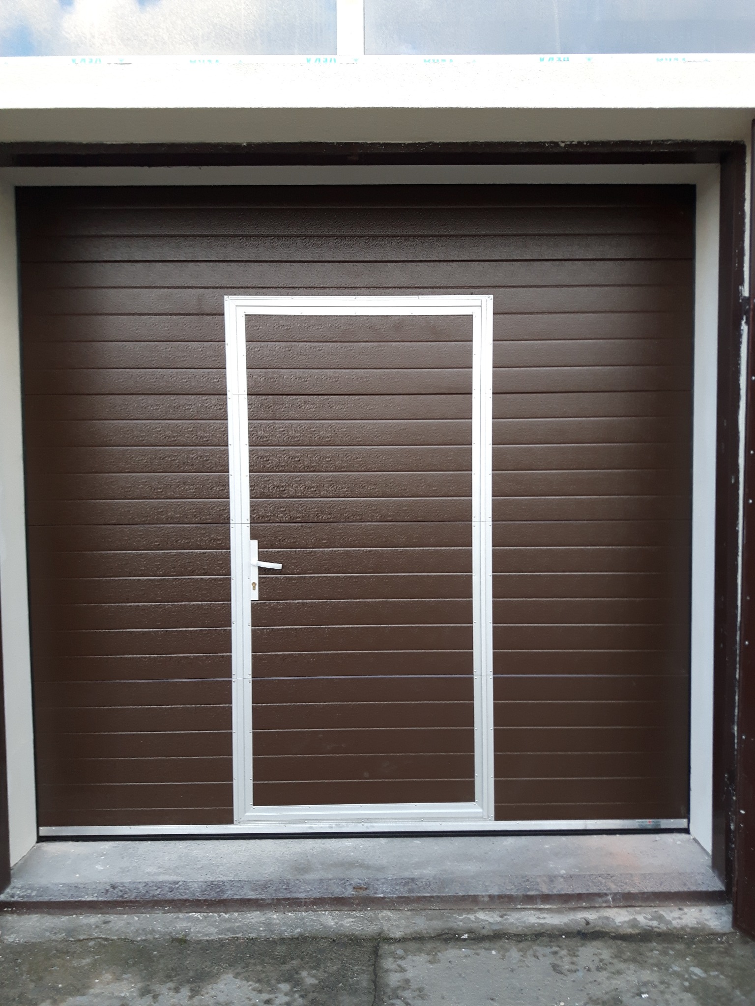 Zewnętrzna, brązowa brama garażowa segmentowa z wbudowanymi drzwiami wejściowymi z białym obramowaniem i klamką, widoczna od frontu.