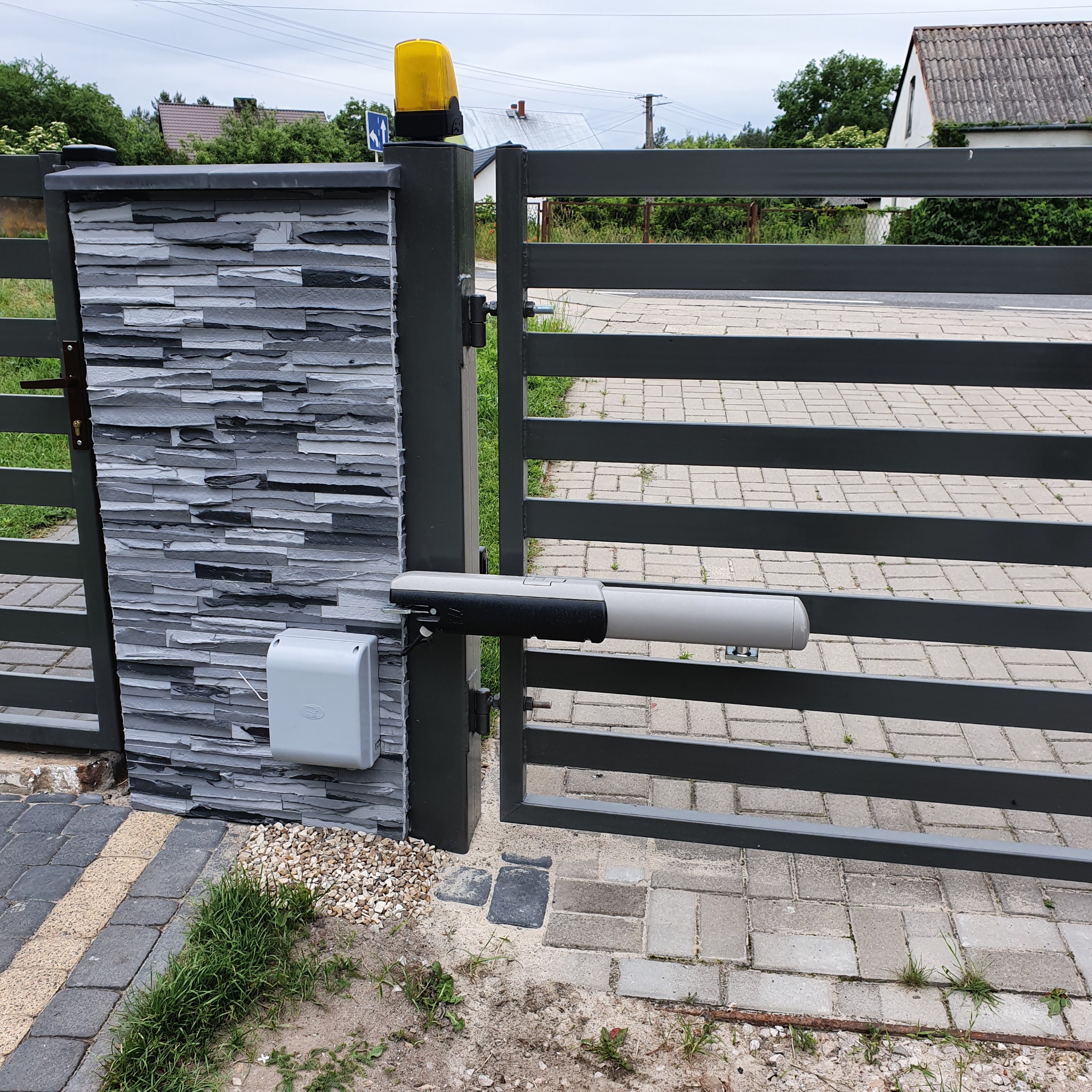 Szara brama skrzydłowa z zamontowanym napędem, widoczna lampa ostrzegawcza na słupku i obudowa sterowania na kamiennym murku.