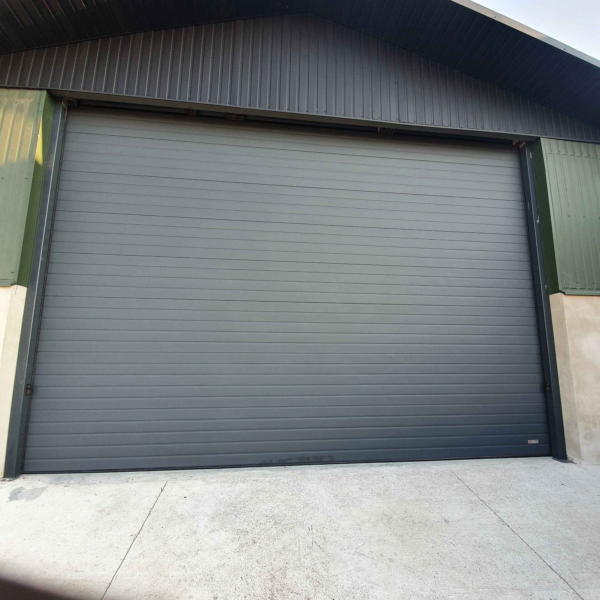 Zamknięta, szara brama segmentowa garażowa z poziomymi przetłoczeniami, widoczna od zewnątrz, z betonową posadzką i fragmentem budynku z zieloną blachą trapezową.