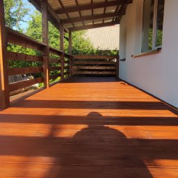 DRY HOUSE PAWEŁ SUSZYCKI - Zadaszony taras drewniany z brązowych desek, widok z dołu, cień osoby na podłodze, drewniana balustrada i słupy, biała ściana budynku w tle.