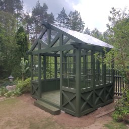DRY HOUSE PAWEŁ SUSZYCKI - Zadaszona altana ogrodowa w odcieniach zieleni, z przezroczystym dachem, otoczona bujną roślinnością. Widoczne detale konstrukcyjne i naturalne oświetlenie.