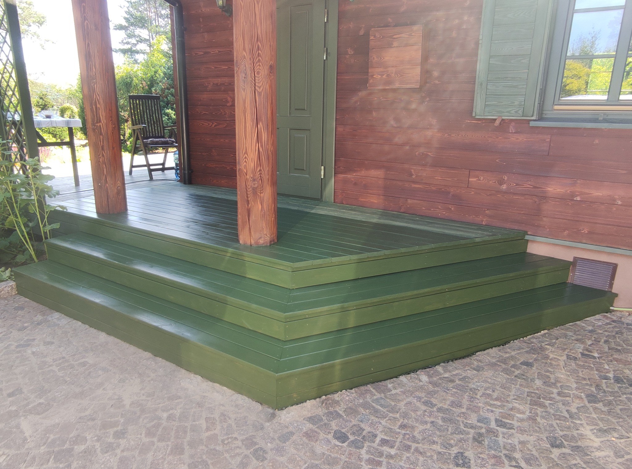 Zielone, drewniane schody wejściowe do domu z brązowymi ścianami i kolumnami. Widoczne okno z okiennicami i kamienna nawierzchnia.