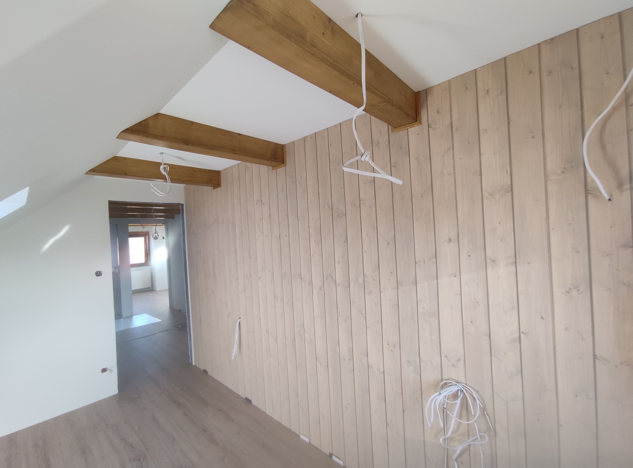 Poddasze z drewnianą boazerią w pionie, bejcowanymi belkami stropowymi i kablami wystającymi ze ściany. Widoczna podłoga i fragment sąsiedniego pomieszczenia.