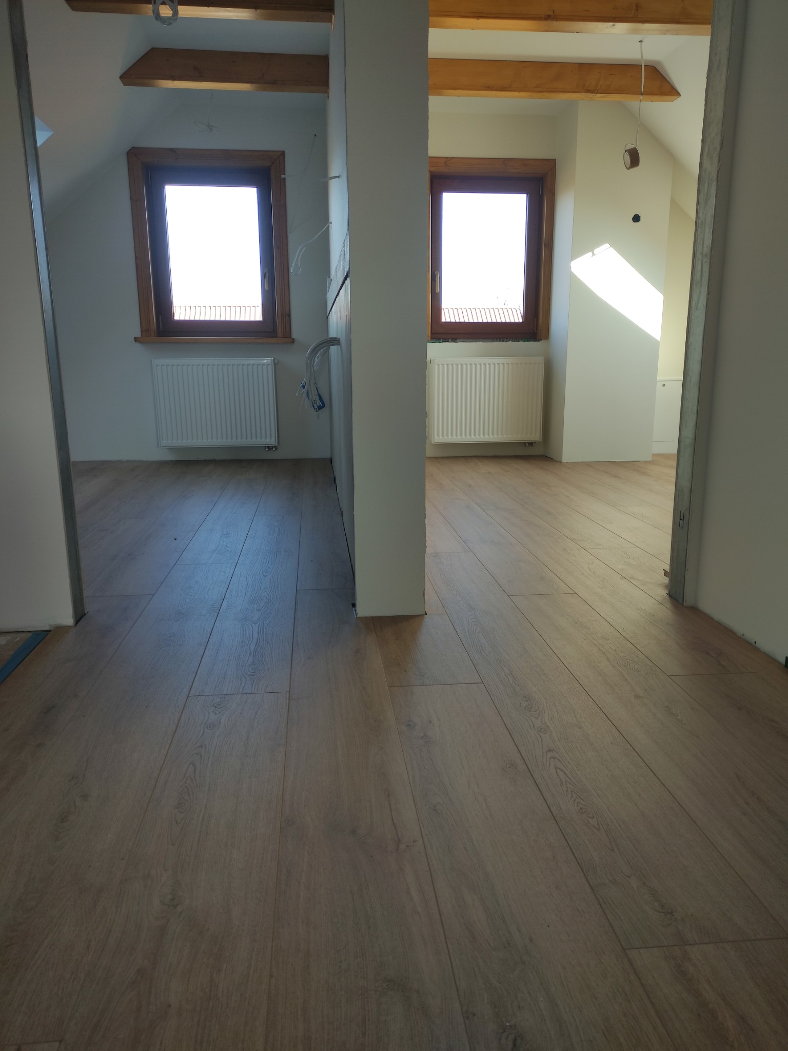 Jasne panele podłogowe w nowym poddaszu z oknami i grzejnikami. Widok przez futrynę, surowe wykończenie ścian, drewniane belki stropowe.