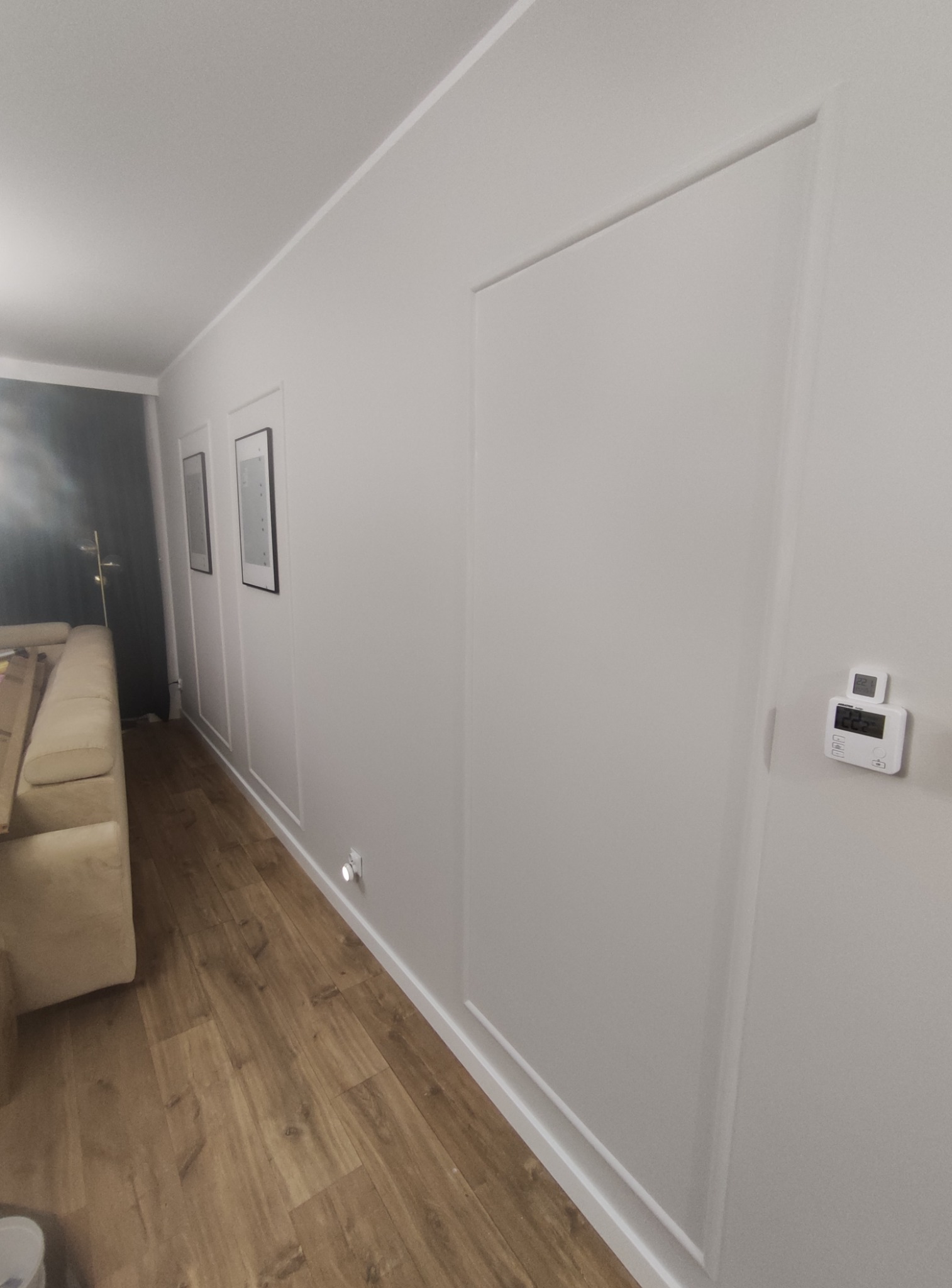 Nowoczesne wnętrze z drewnianą podłogą, białe ściany z dekoracyjnymi panelami i ukrytymi drzwiami. Minimalistyczny design z oświetleniem punktowym i termostatem.