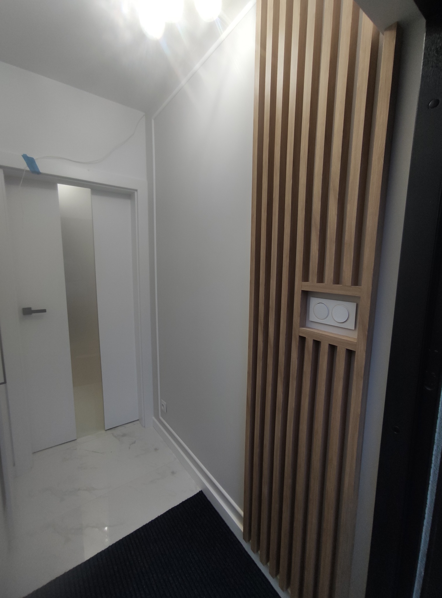 Nowoczesny przedpokój z drewnianą ścianką działową i wbudowanym włącznikiem światła. Białe ściany, marmurowa podłoga i minimalistyczne drzwi.