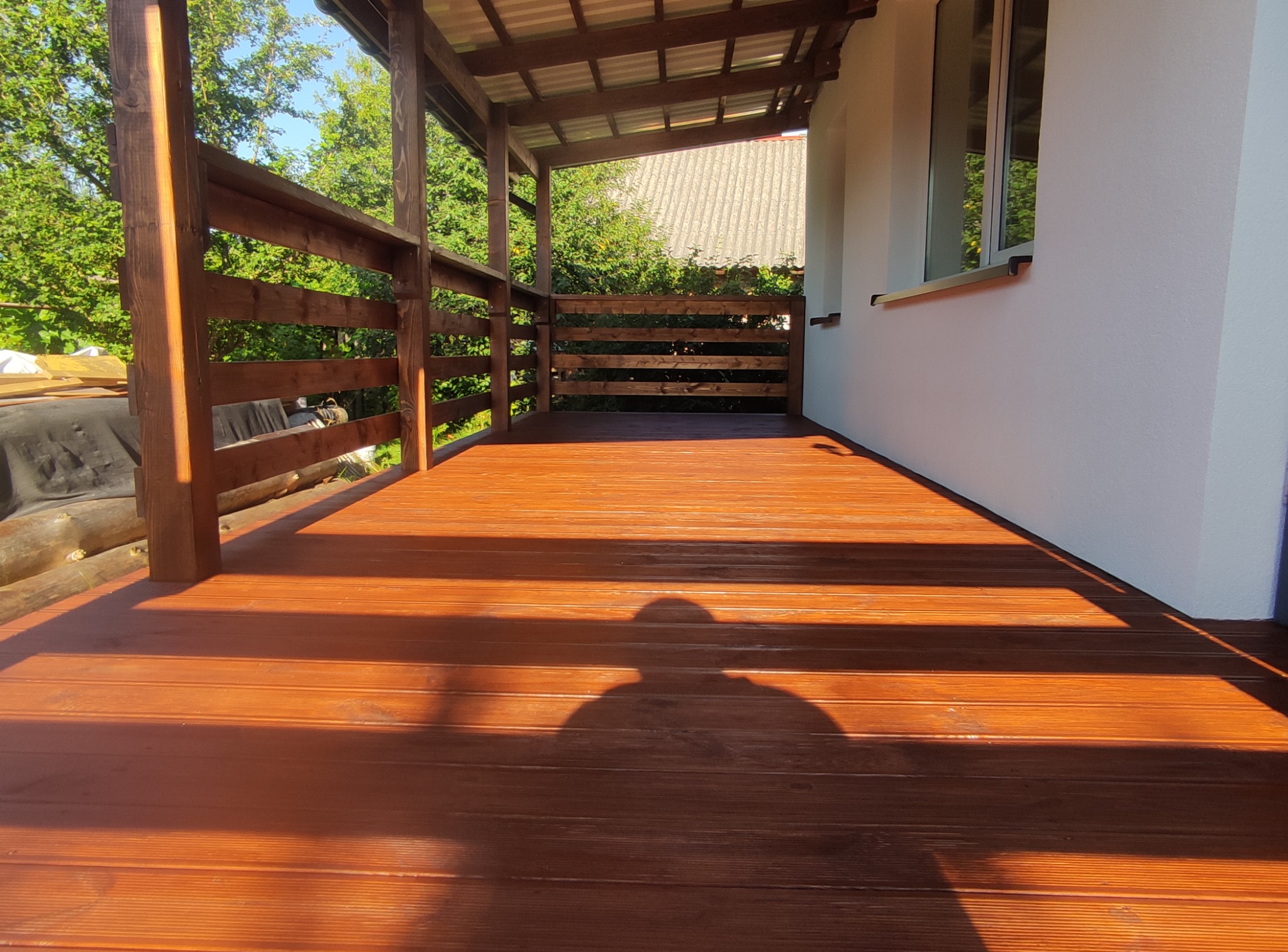 Zadaszony taras drewniany z brązowych desek, widok z dołu, cień osoby na podłodze, drewniana balustrada i słupy, biała ściana budynku w tle.