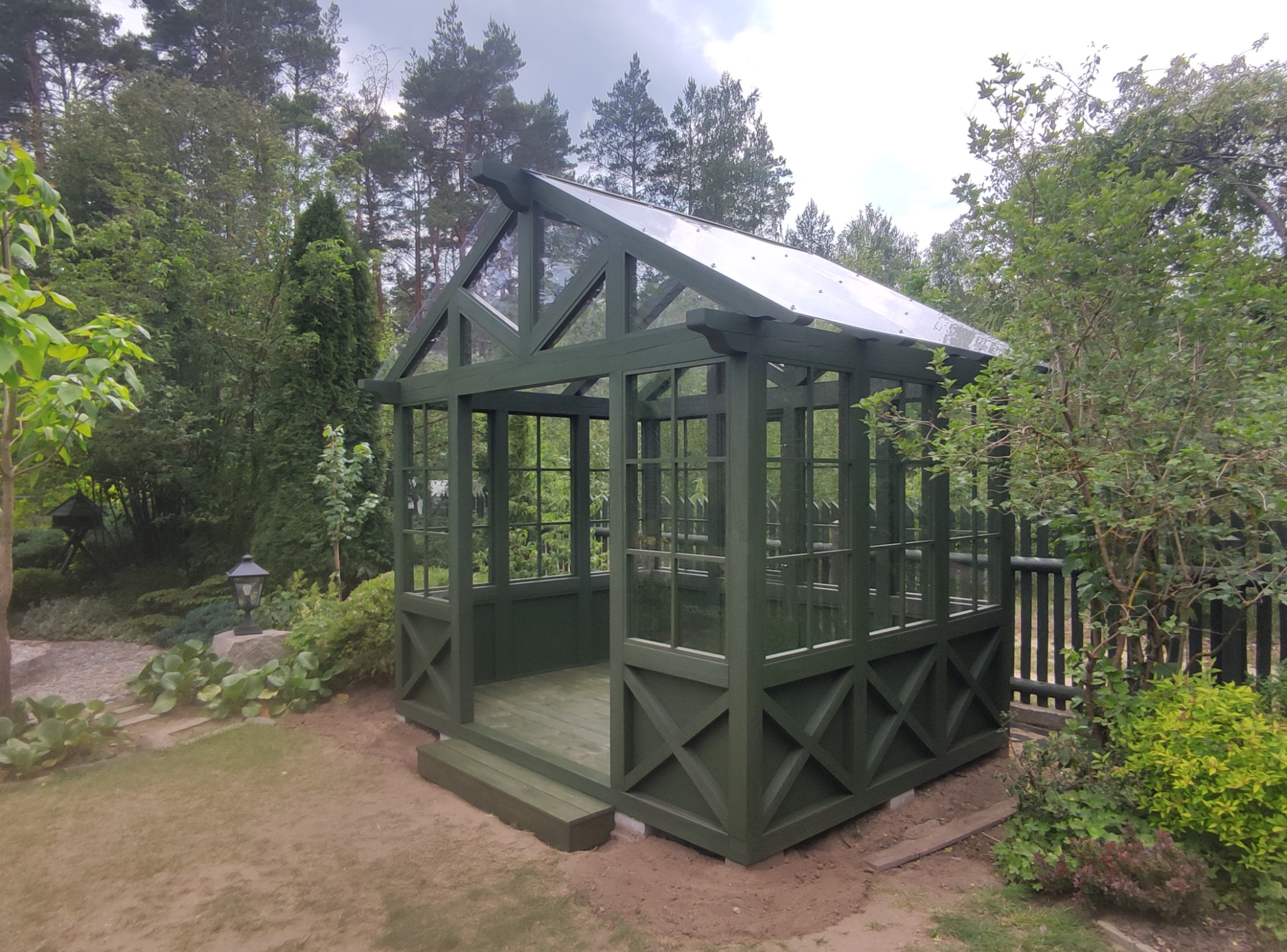 Zadaszona altana ogrodowa w odcieniach zieleni, z przezroczystym dachem, otoczona bujną roślinnością. Widoczne detale konstrukcyjne i naturalne oświetlenie.