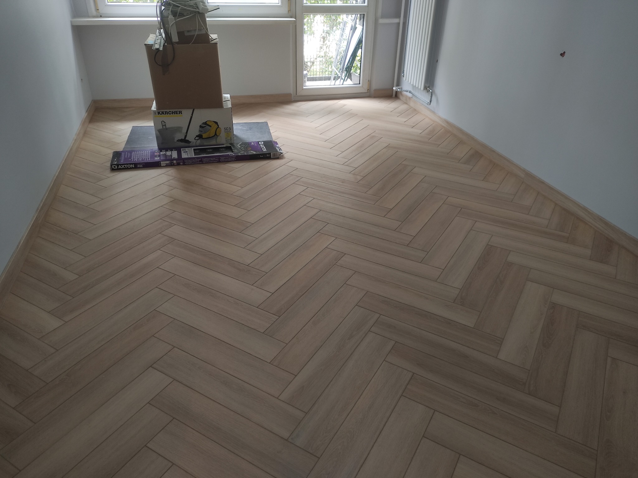 Jasne panele ułożone w jodełkę w pustym pokoju z oknem. Na podłodze kartony i opakowania po sprzęcie. Widoczne listwy przypodłogowe i grzejnik.