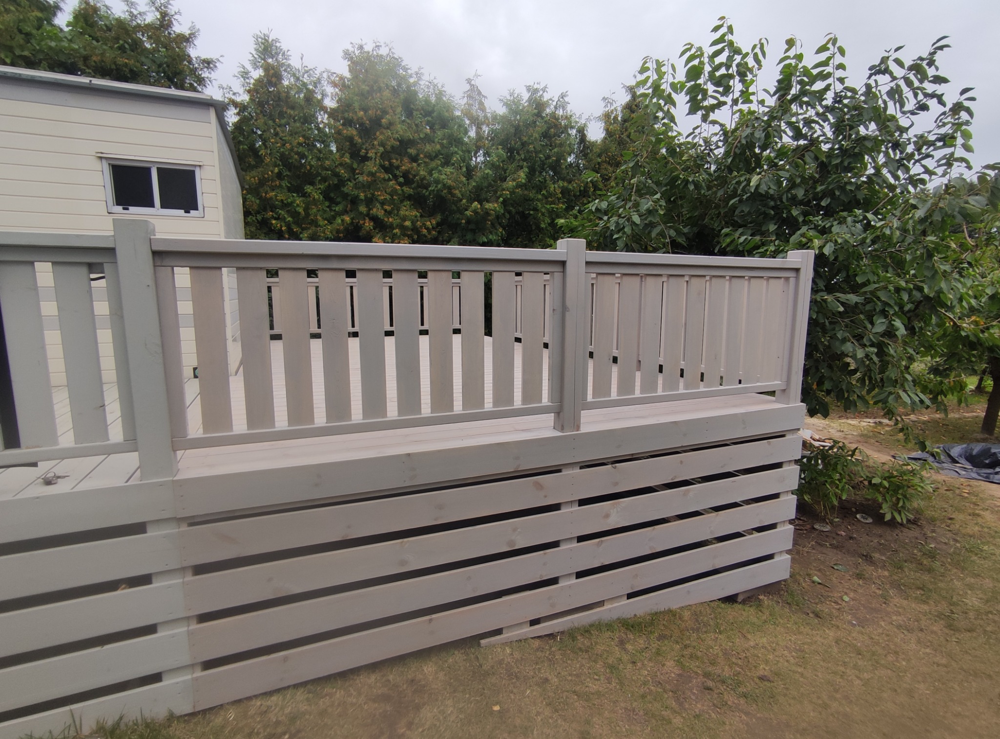 Jasnoszary taras drewniany z balustradą w Białymstoku, widok z poziomu trawnika. W tle zieleń drzew i fragment budynku z oknem. Drewniana konstrukcja.