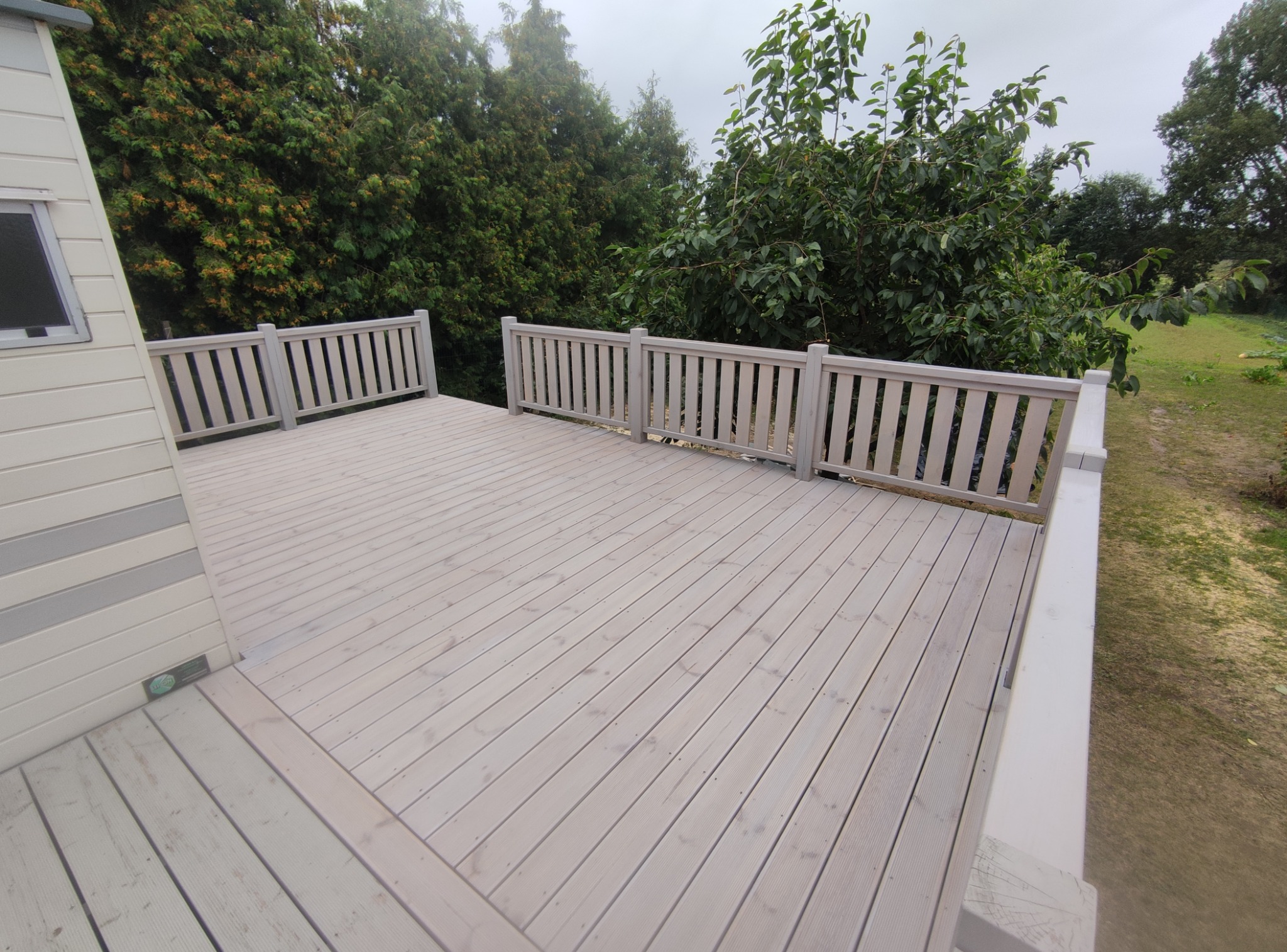 Jasny, drewniany taras z balustradą, otoczony zielenią drzew i krzewów. Widok z góry ukazuje deski ułożone równolegle, tworząc przestronną i estetyczną powierzchnię.