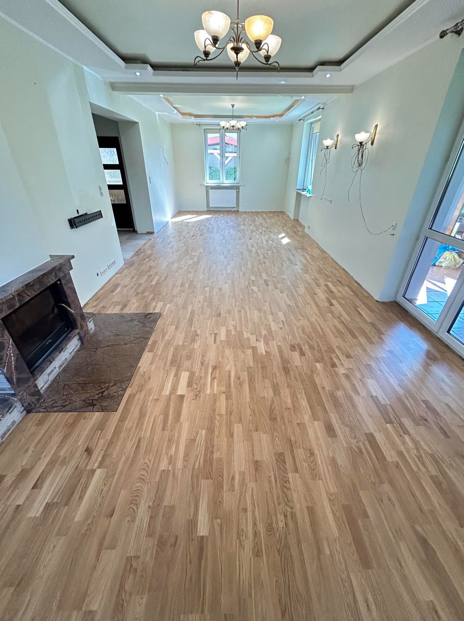 Jasny parkiet ułożony w przestronnym salonie z kominkiem i dużymi oknami. Widoczne niedokończone instalacje elektryczne na ścianach.