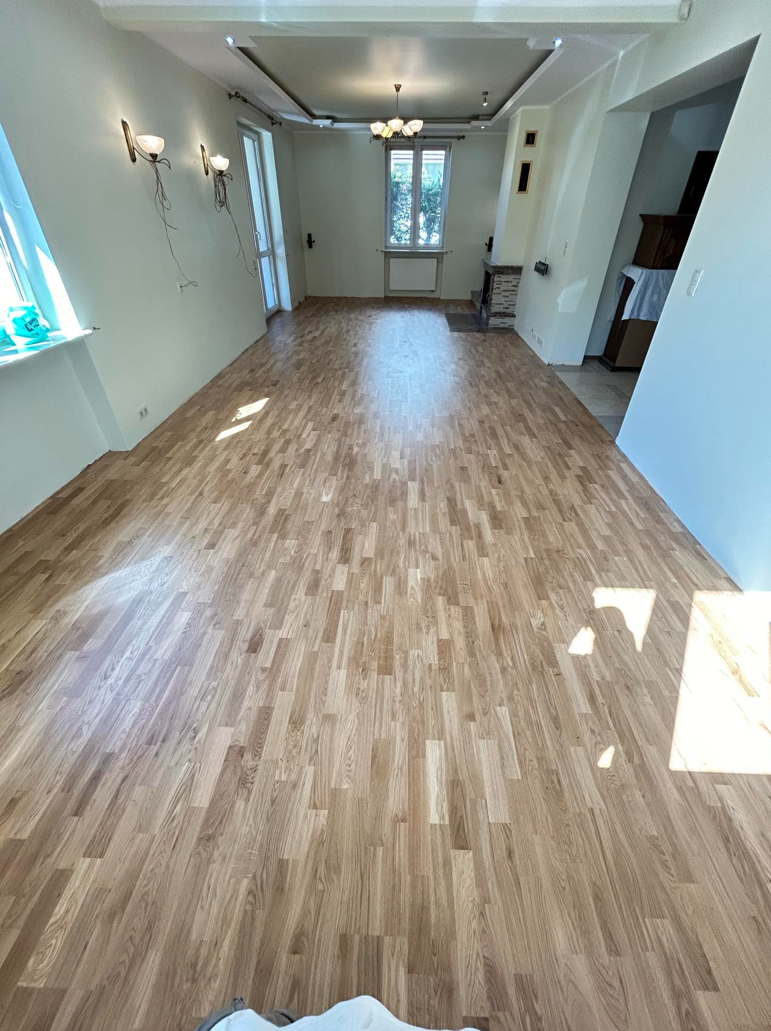 Jasne panele podłogowe ułożone w przestronnym, odnowionym salonie z oknem i kominkiem. Świeżo pomalowane ściany i nowoczesne oświetlenie.