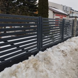 Bramax Urszula Janocińska - Nowoczesne, poziome ogrodzenie panelowe w kolorze grafitowym, z widocznym pracownikiem podczas montażu. Tło stanowi budynek Ochotniczej Straży Pożarnej w zimowej scenerii.