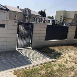 Bramax Urszula Janocińska - Nowoczesne ogrodzenie panelowe z bramką furtką, z szarej kostki betonowej, z widocznym domem w tle. Furtka rzuca cień na kostkę. Widok na ogrodzenie z zewnątrz posesji.