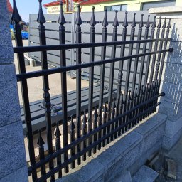 Bramax Urszula Janocińska - Czarne, metalowe ogrodzenie kute z ozdobnymi grotami, zamontowane na betonowej podmurówce. W tle widoczny budynek z czerwonym dachem i panelami słonecznymi.