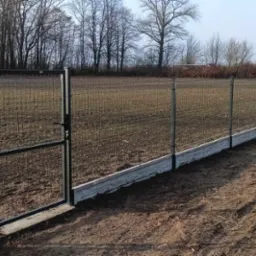 Panelowe ogrodzenie z bramą wjazdową, osadzone na betonowej podmurówce. Widoczne metalowe słupki i siatka ogrodzeniowa. Tło stanowią drzewa i pole.
