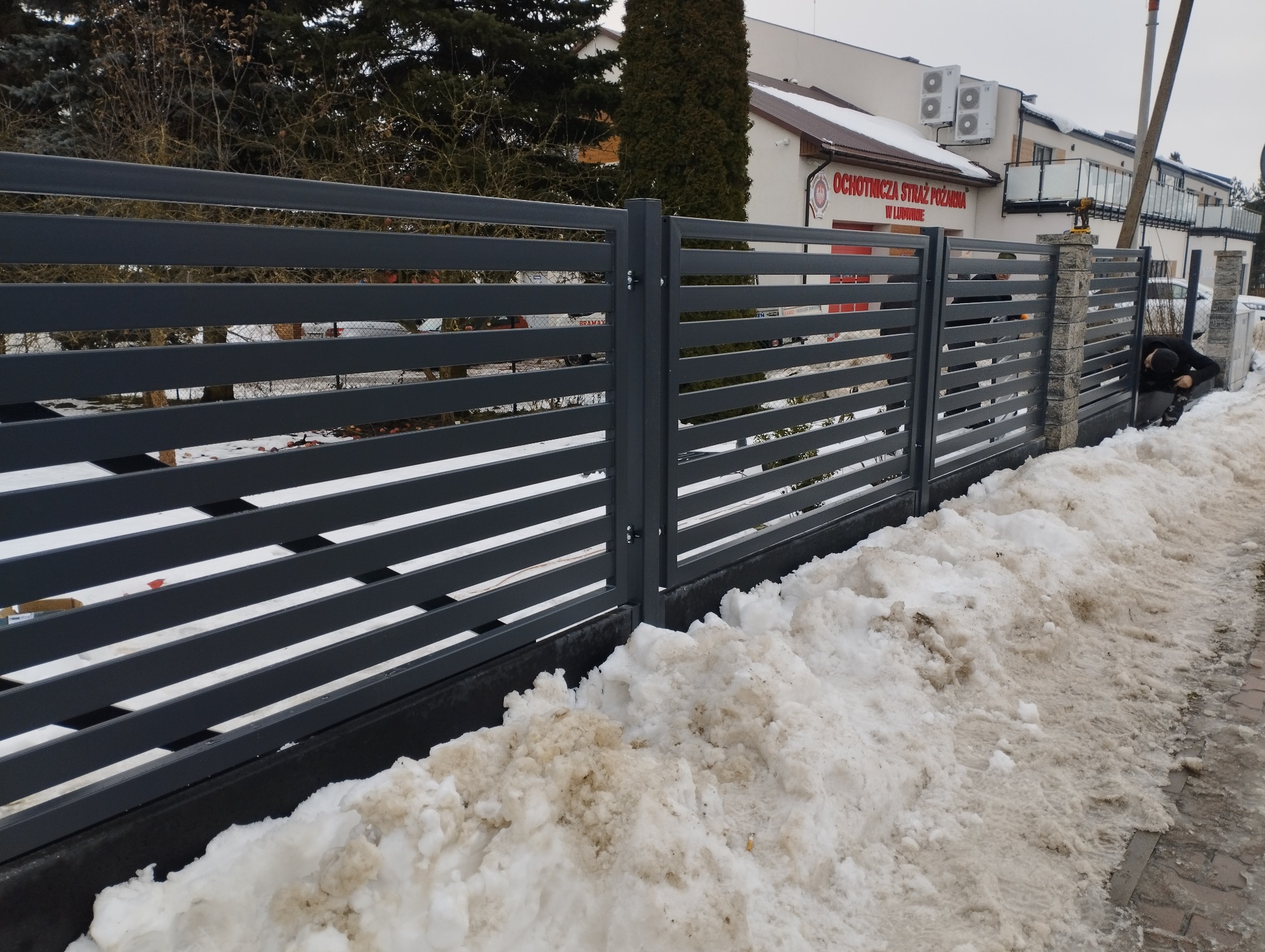 Nowoczesne, poziome ogrodzenie panelowe w kolorze grafitowym, z widocznym pracownikiem podczas montażu. Tło stanowi budynek Ochotniczej Straży Pożarnej w zimowej scenerii.