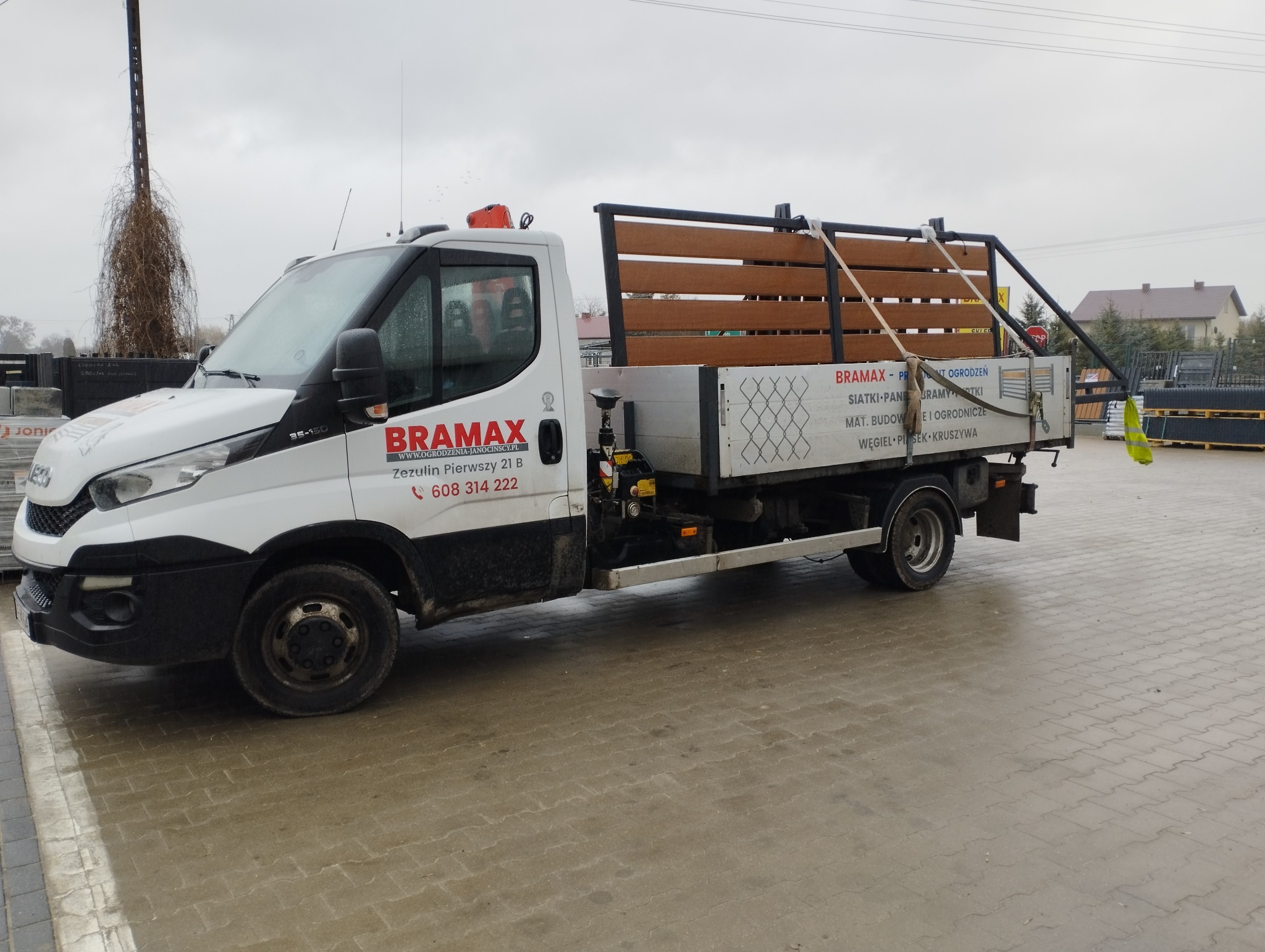 Ciężarówka firmy Bramax z panelami ogrodzeniowymi na pace, zaparkowana na placu. Widoczne ogrodzenia w tle i dane kontaktowe na pojeździe.