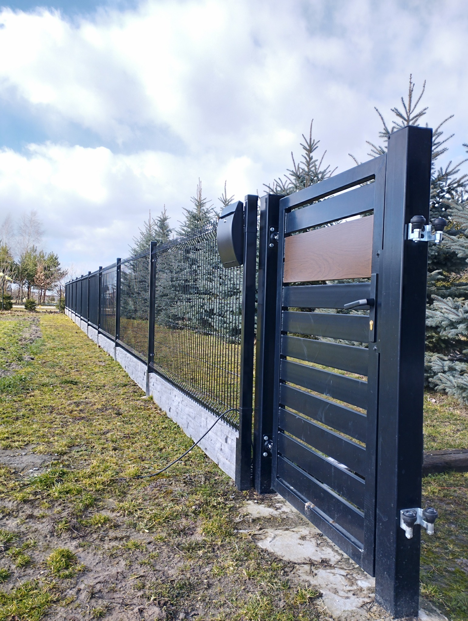 Nowoczesne ogrodzenie panelowe z bramą wjazdową, czarne słupki, podmurówka z imitacji kamienia, skrzynka na listy. Widok z boku, trawnik i drzewa w tle.