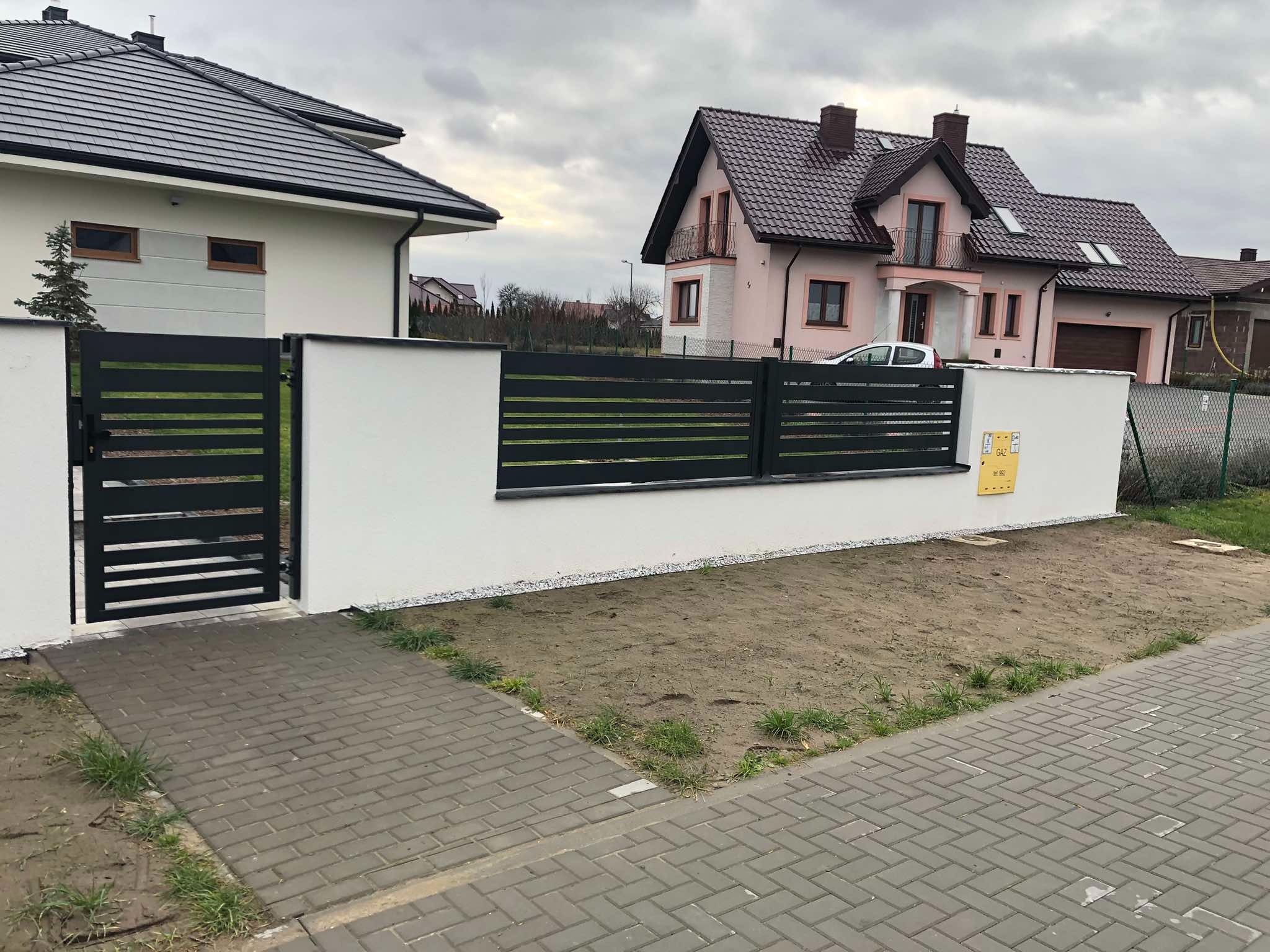 Nowoczesne, poziome ogrodzenie panelowe w kolorze antracytowym, zamontowane na białym murku. W tle widoczne dwa domy jednorodzinne i pochmurne niebo.
