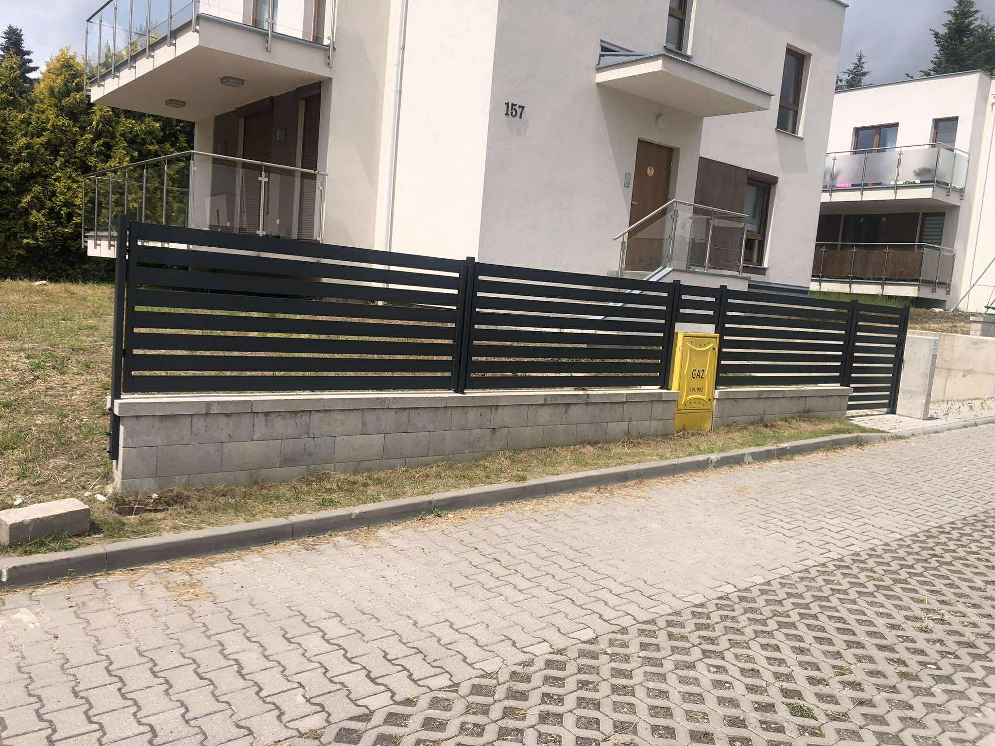 Nowoczesne, poziome ogrodzenie panelowe w kolorze antracytowym, zamontowane na betonowej podmurówce przed budynkiem mieszkalnym o jasnej elewacji.