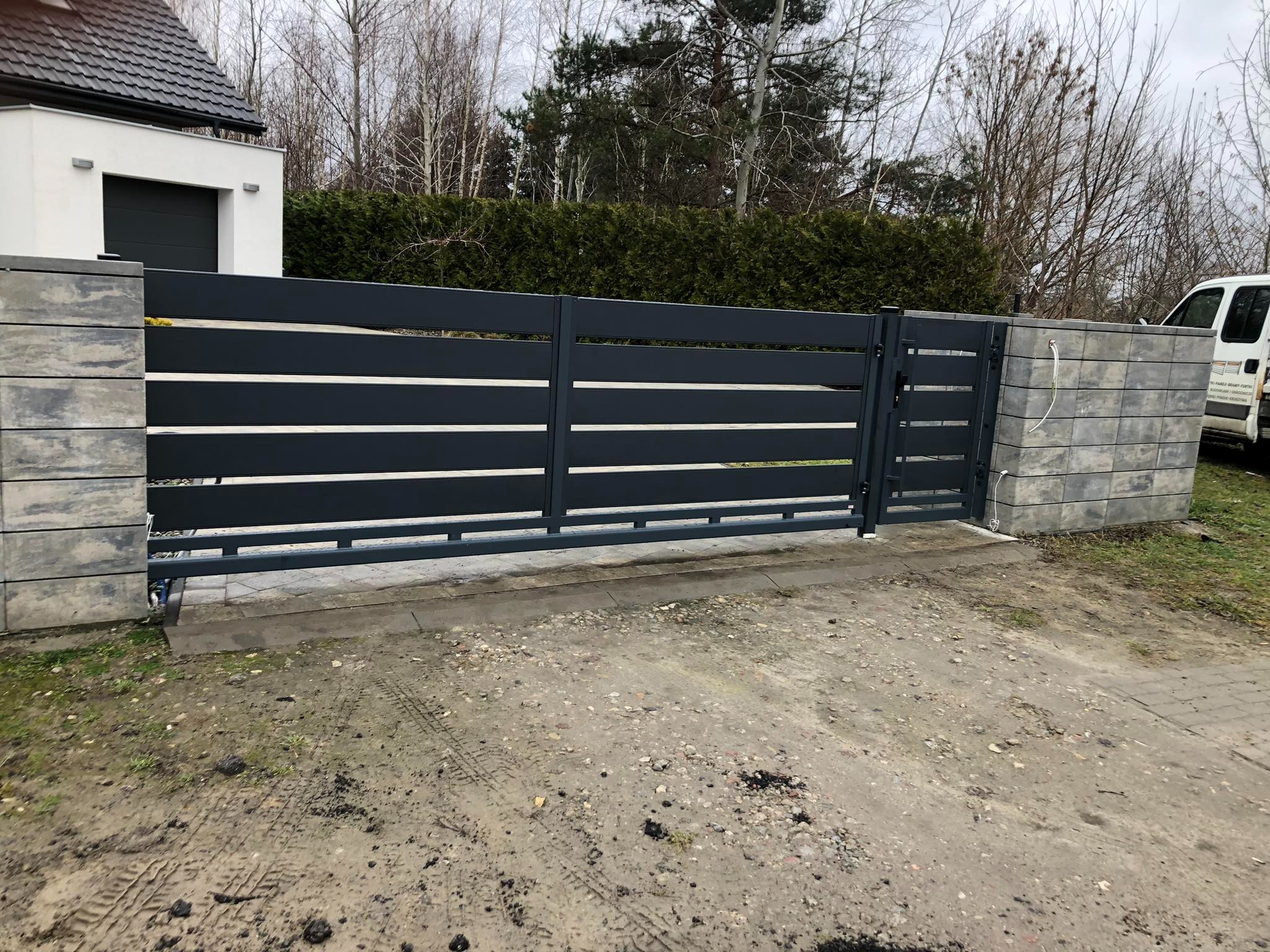 Nowoczesna, pozioma brama wjazdowa w kolorze grafitowym, osadzona na betonowych słupkach. Widoczny fragment domu z garażem i otoczenie z drzewami.